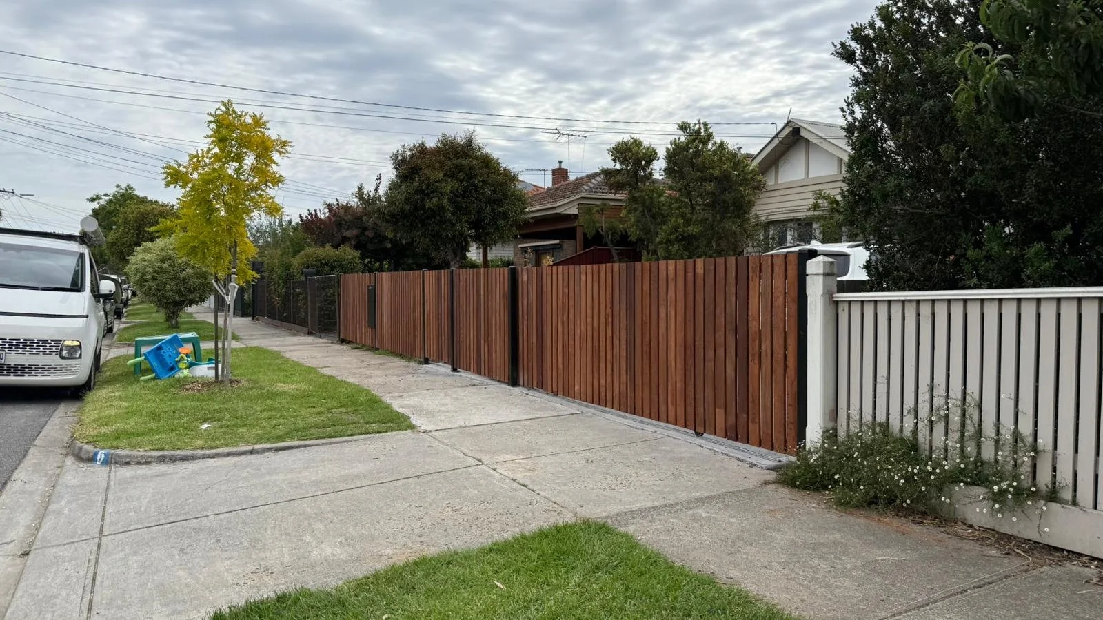 A sidewalk next to a residential street with a row of trees, parked cars, and a wooden fence with different sections. There is a small grassy area with a tree and some children's toys. The sky is overcast.