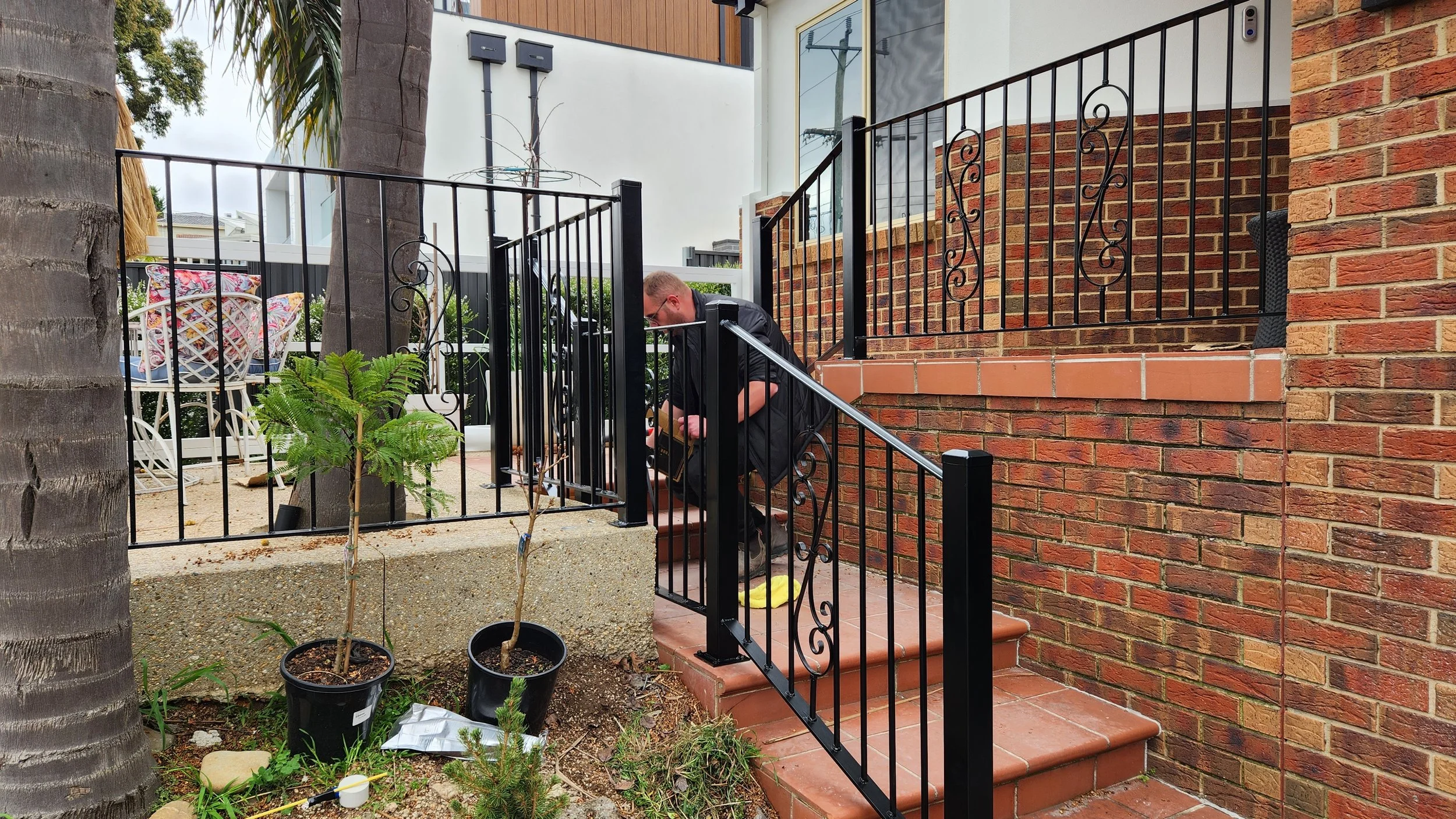 A person crouching on the stairs outside a building, working on a black metal railing. There are potted plants and gardening tools nearby, with a brick and white wall in the background.