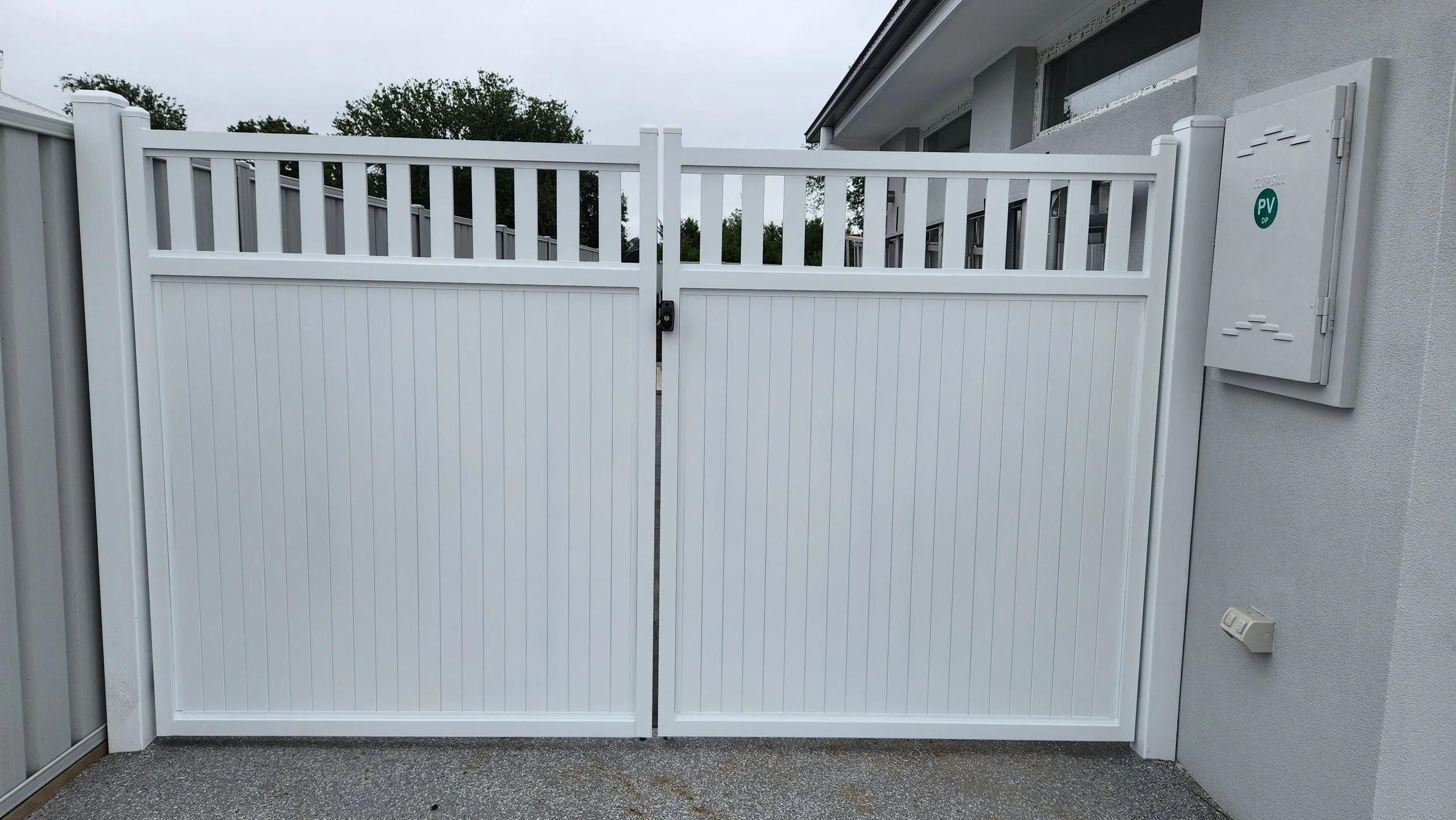 White double gate with vertical panel design in front of a residential house, with utility panels on the wall to the right.