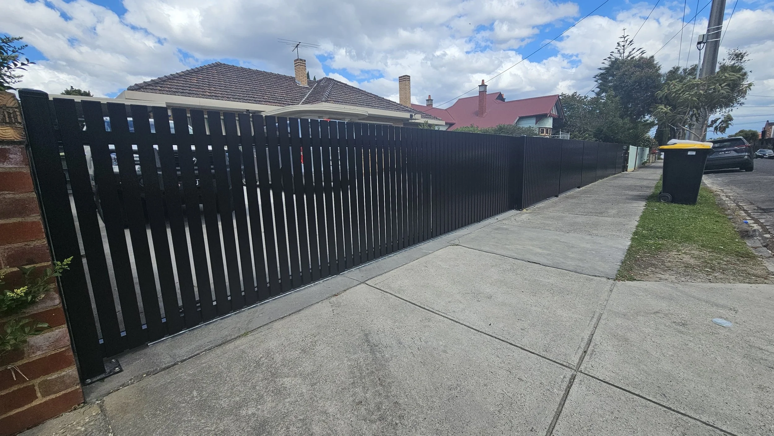 Black sliding fence gate along sidewalk with houses and parked cars in the background under partly cloudy sky.