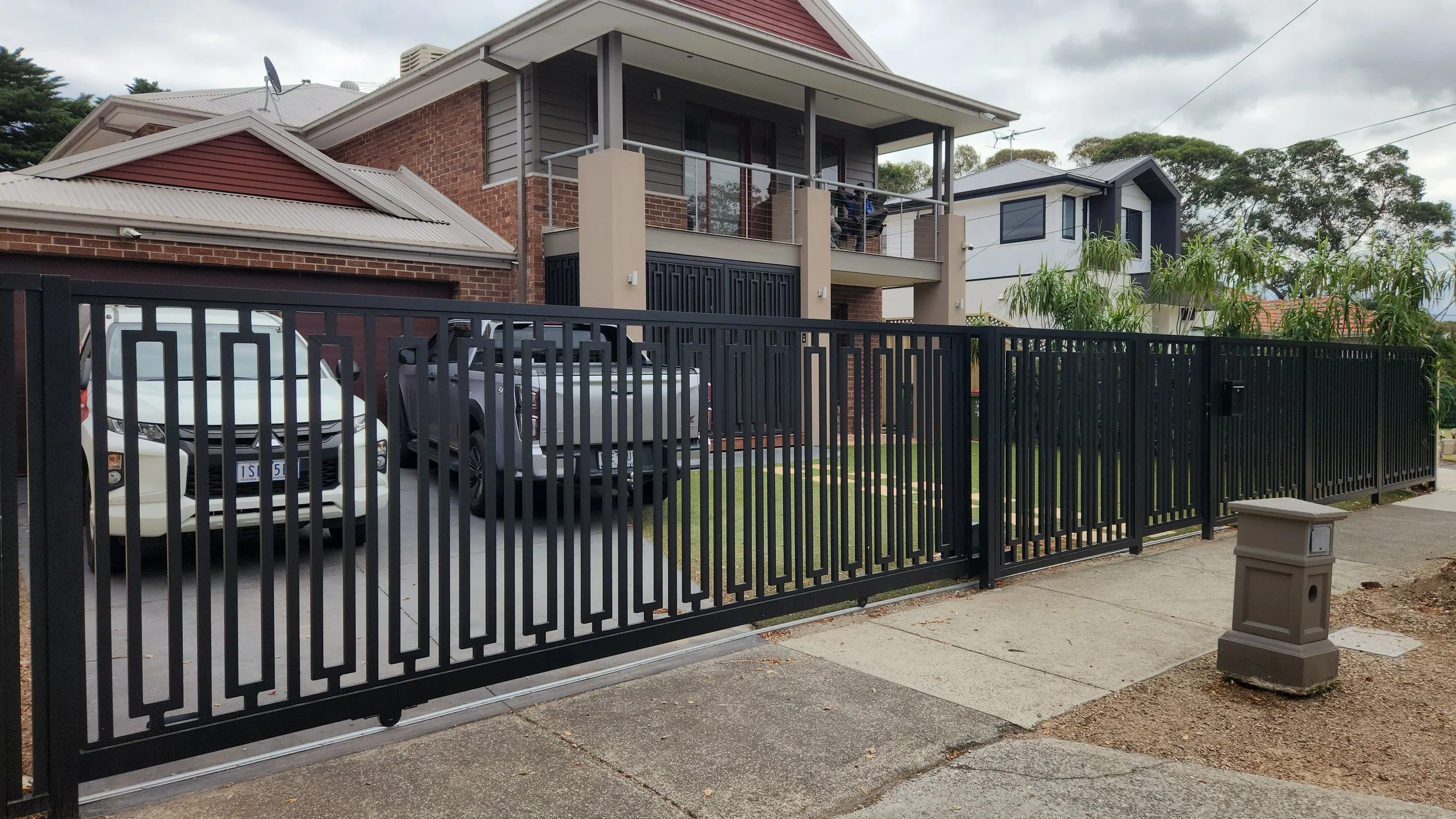 Modern residential house with a black metal fence, driveway with two parked cars, and a two-story balcony. Neighboring houses and trees are visible in the background.