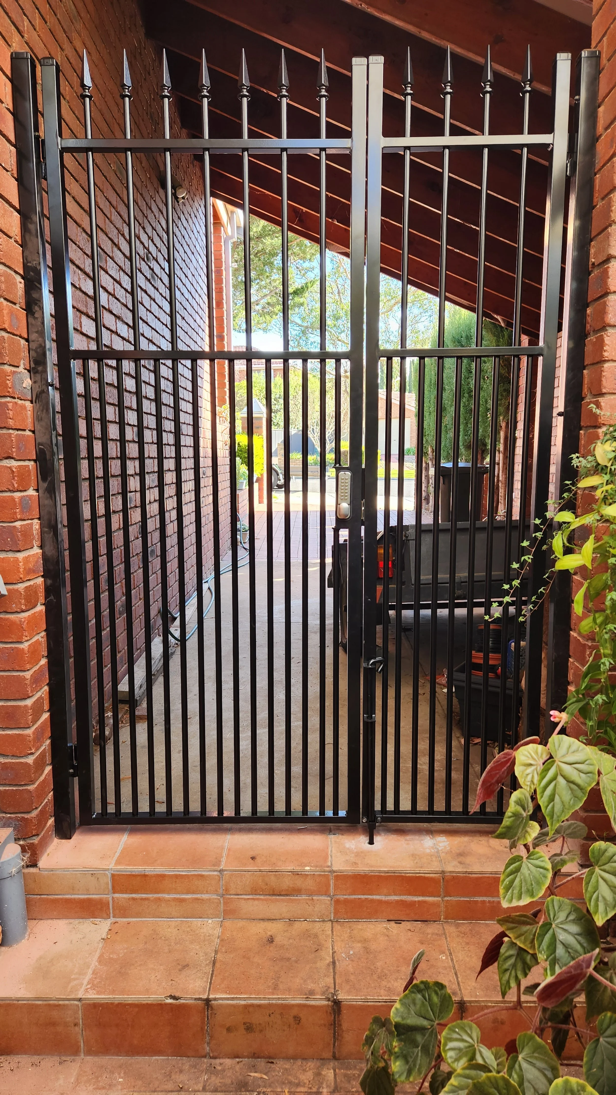 Black metal security gate at entrance of a patio with brick steps and green plants nearby, leading to a sidewalk with houses in the background.
