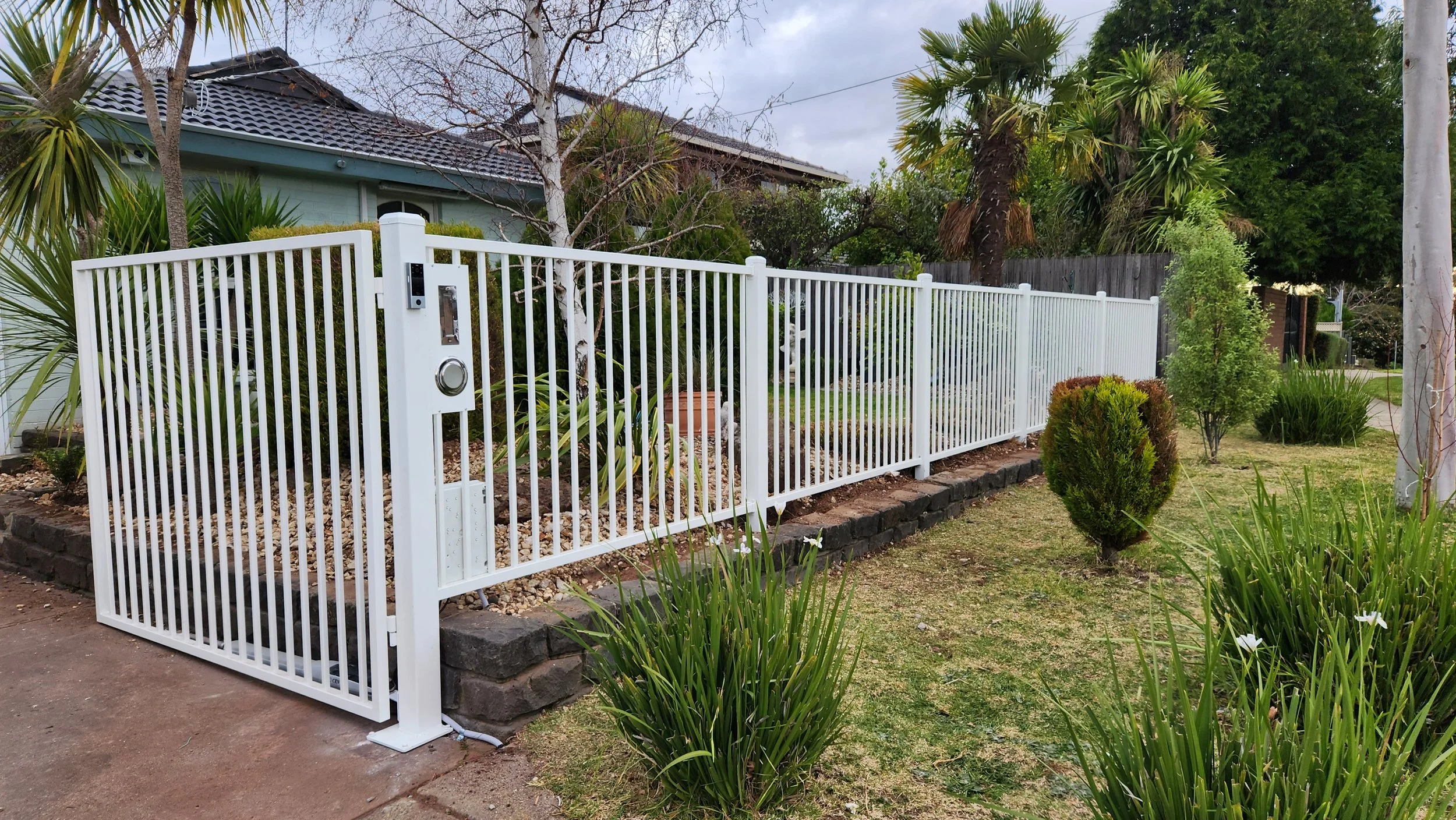 White metal fence with a sliding gate surrounds a landscaped front yard with trees, bushes, and grass in a suburban neighborhood.
