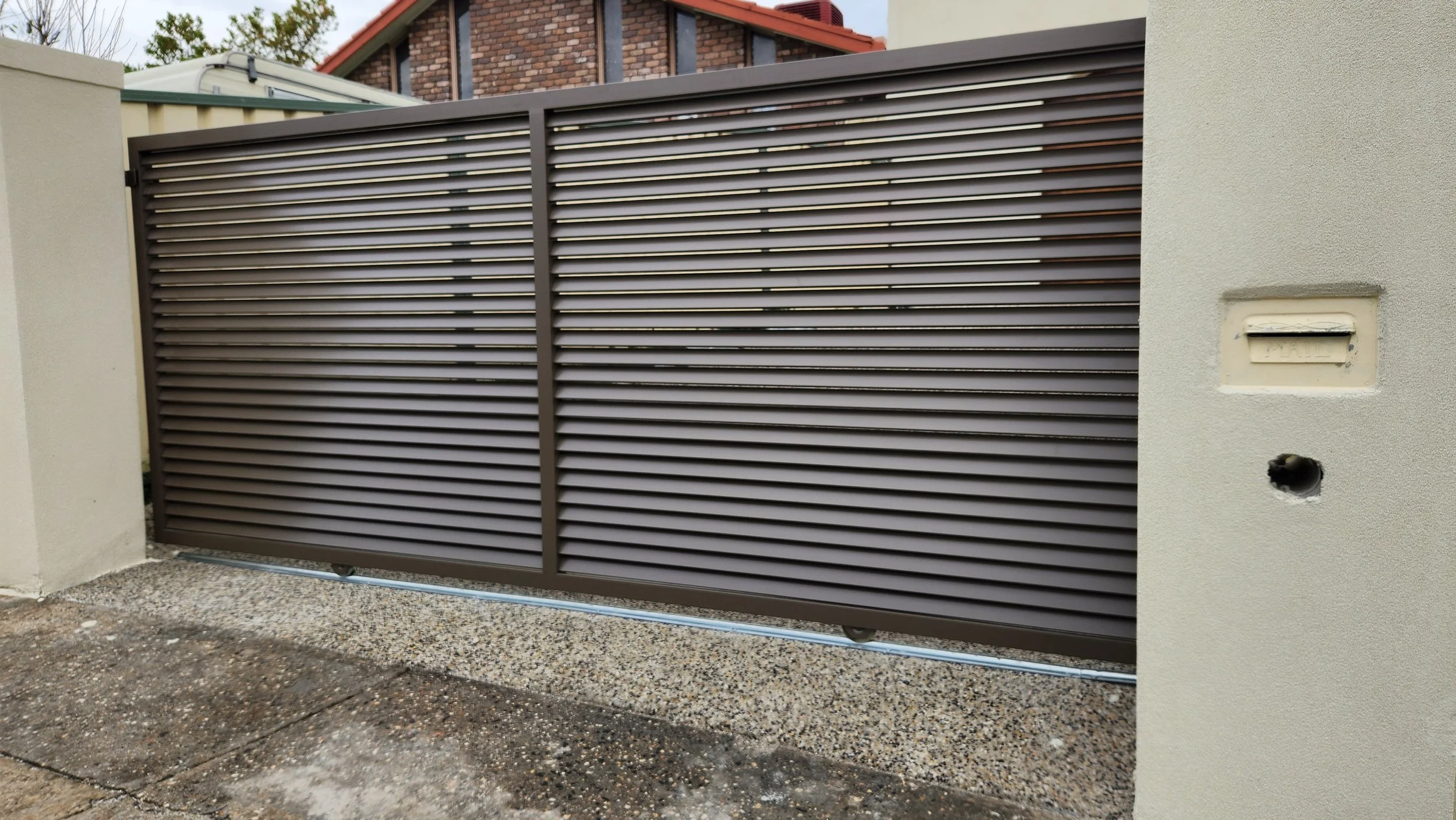 Metal horizontal slat gate with beige stucco walls on each side, driveway surface below, and neighboring houses in background.