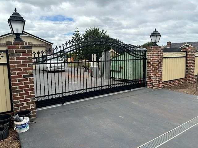 Black wrought iron sliding gate with brick and wooden fence pillars on either side, decorative lamps on top, and a driveway in front.