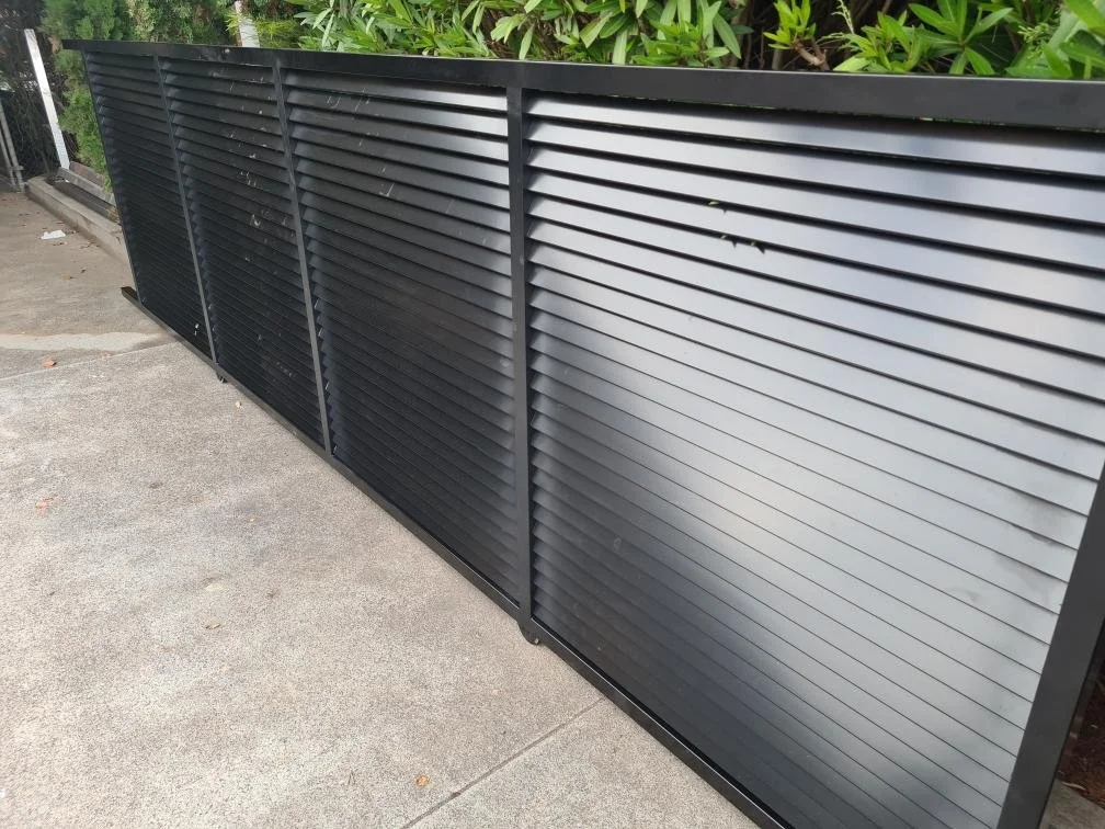 A black metal fence with horizontal slats situated along an outdoor path, with lush green bushes in the background.