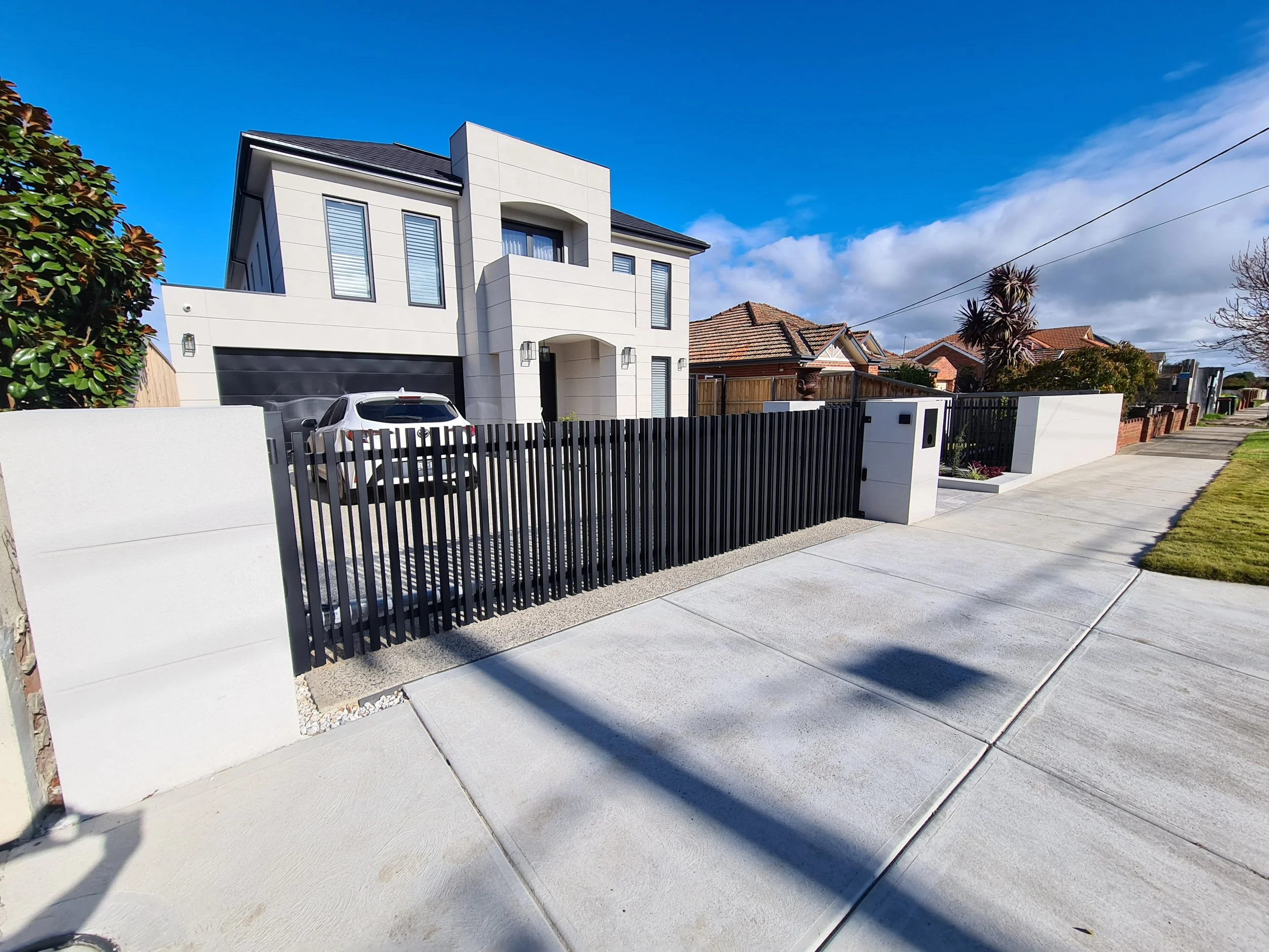 Modern white two-story house with a black garage door, fenced driveway, gray car parked in the driveway, and a well-maintained sidewalk and lawn in front under a blue sky with some clouds.