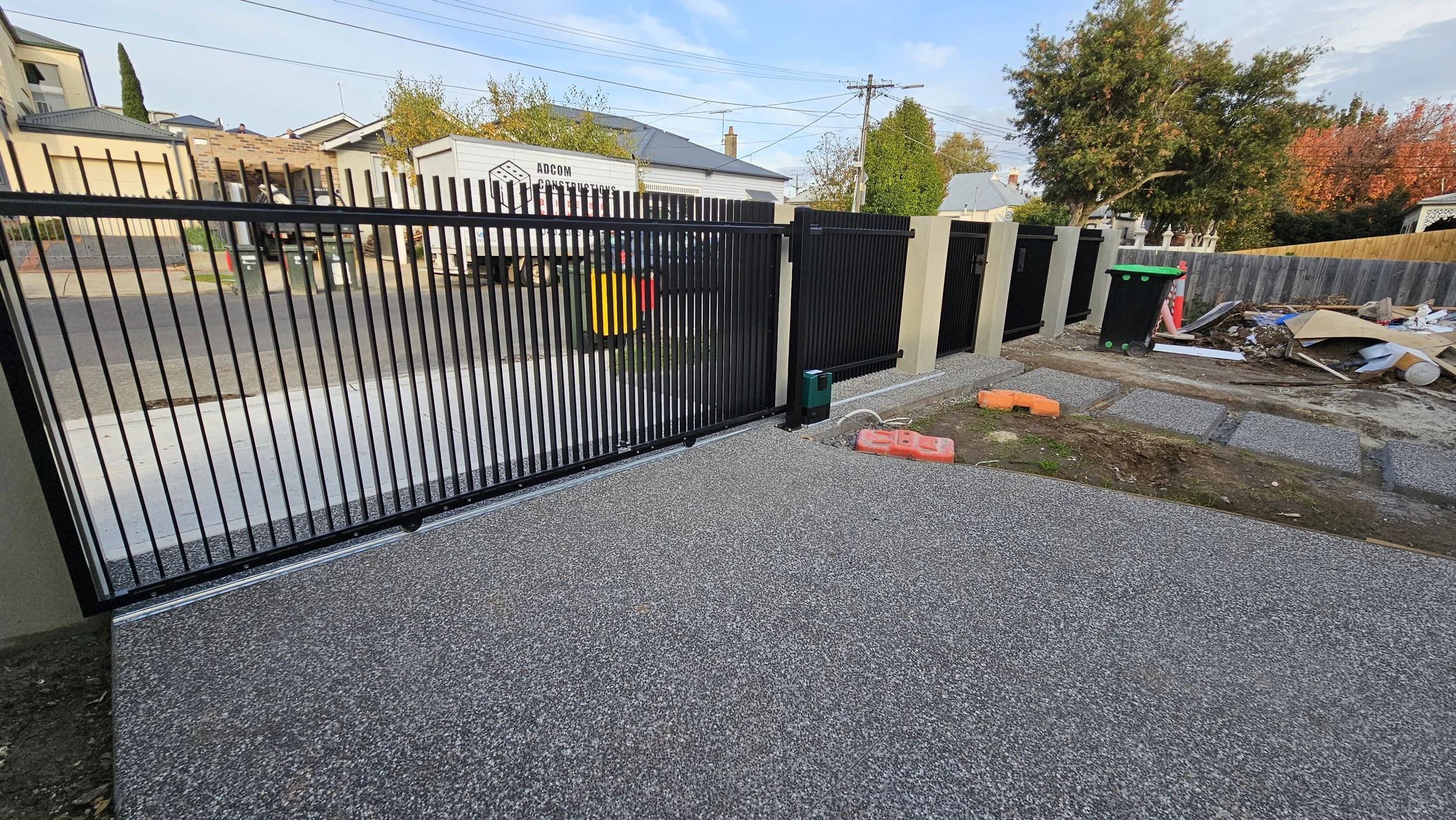 A newly installed black metal driveway gate with a pebble-textured concrete surface in the foreground. In the background, there's a street with parked vehicles, trees, and residential houses, along with construction materials and debris on the right