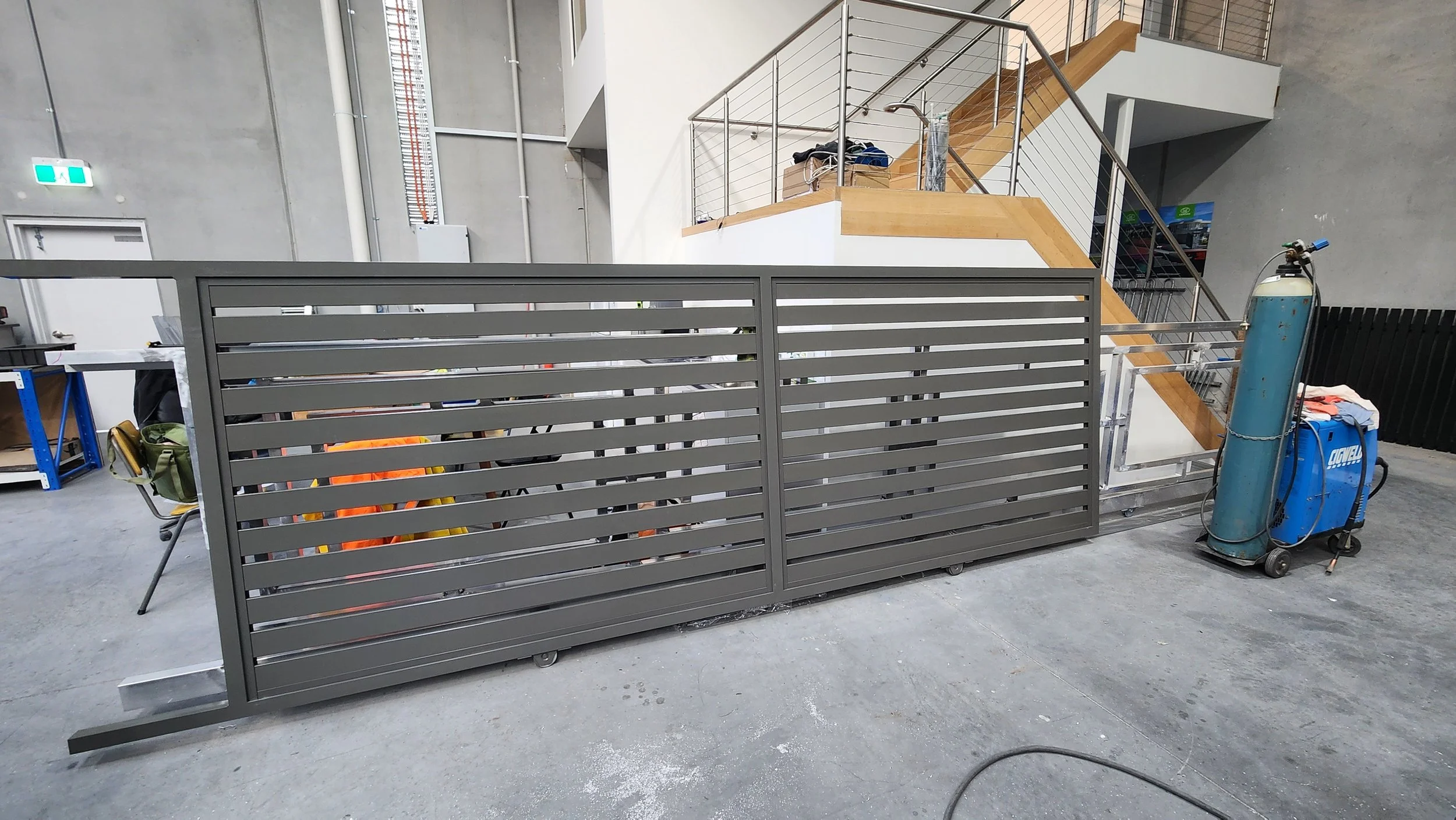 Industrial staircase with safety railings, a large gray metal sliding gate in front, and a welding oxygen tank nearby inside a factory or warehouse