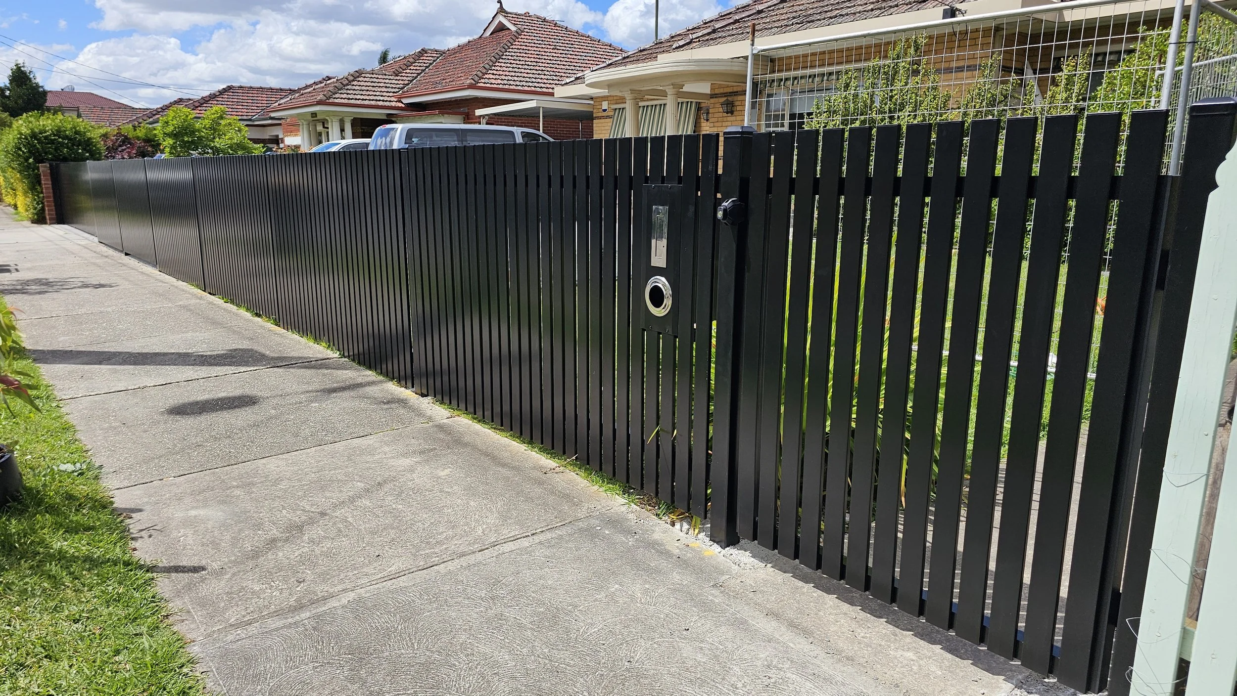A long black metal fence runs along a sidewalk in front of residential houses with red tiled roofs; the fence includes a gate with a handle and a mailbox slot.