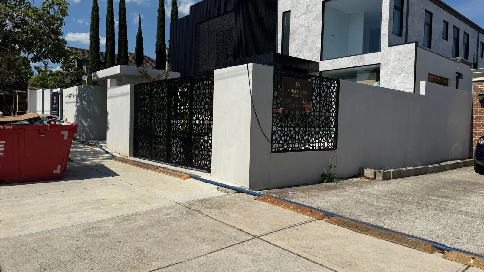 Modern two-story house with gray exterior walls and large windows, enclosed by a concrete wall with a decorative black metal gate, and trees in the background.