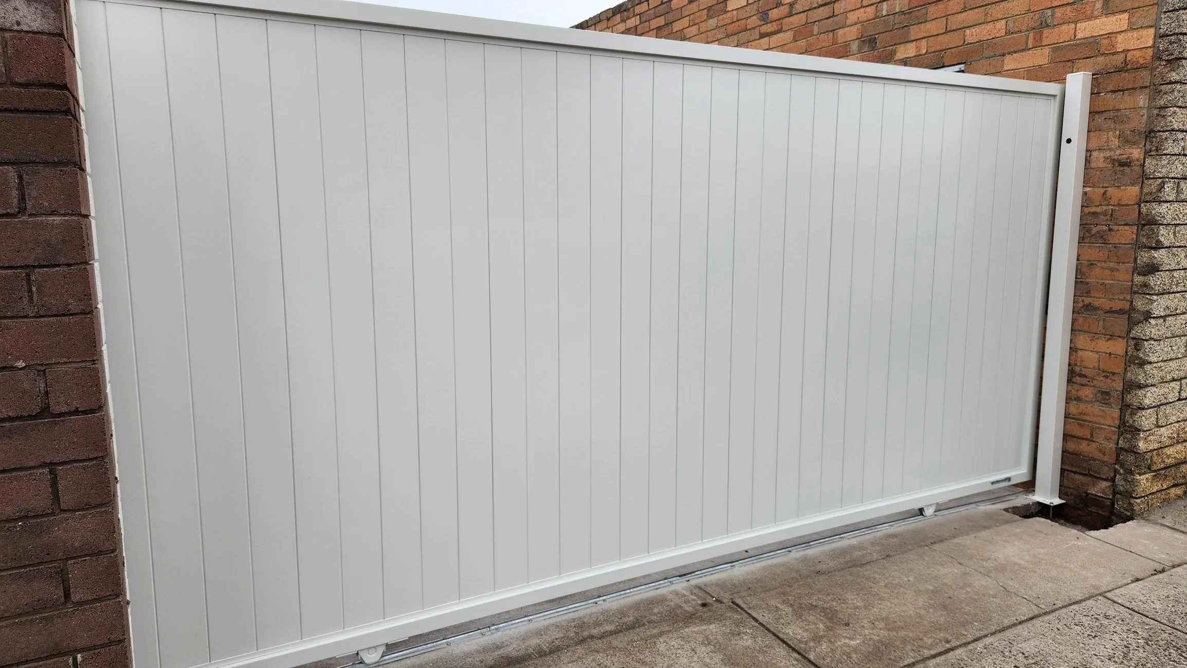 A white metal sliding gate between brick walls on a concrete sidewalk.