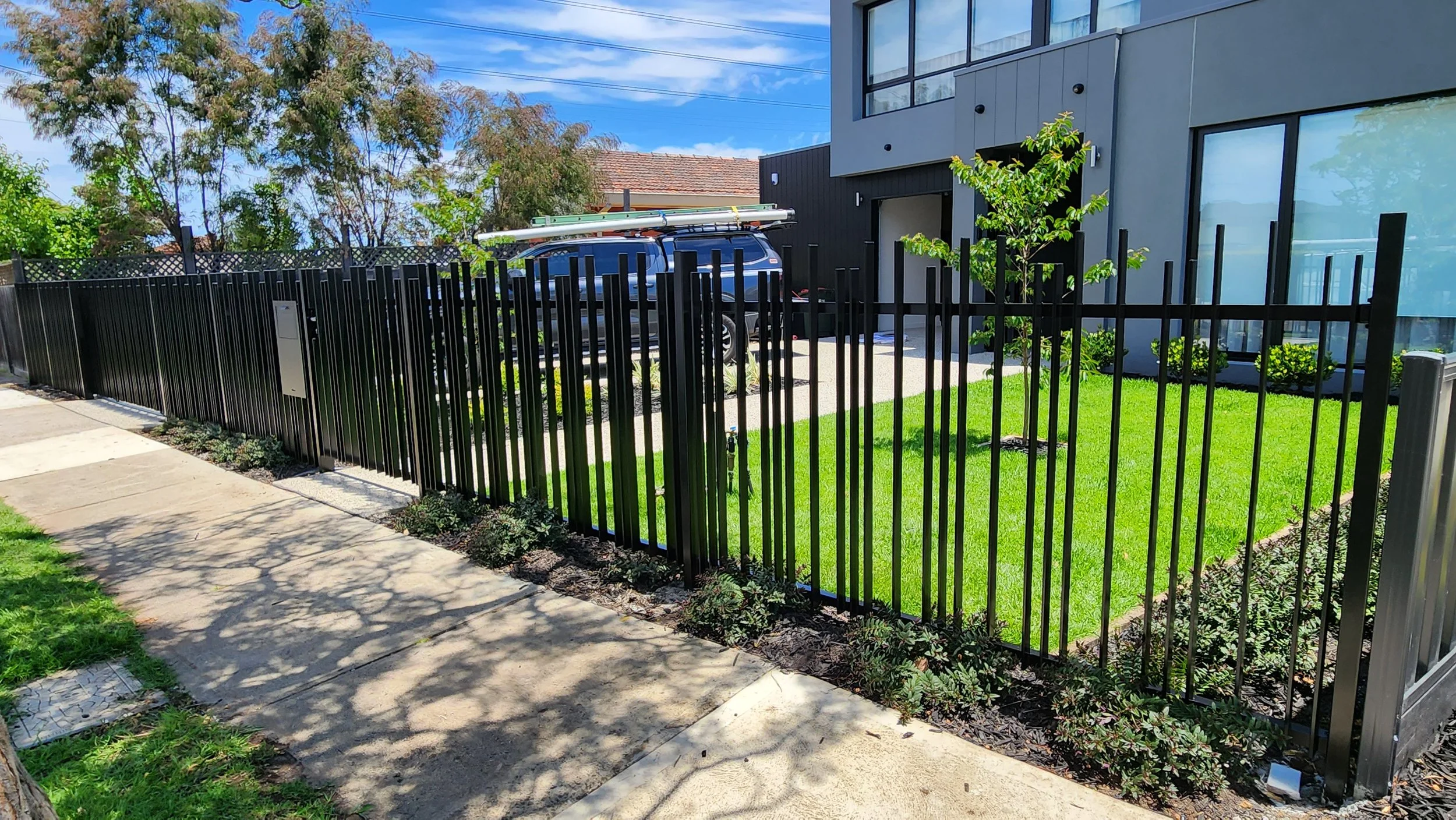 Modern house with black metal fence, green lawn, and sidewalk.