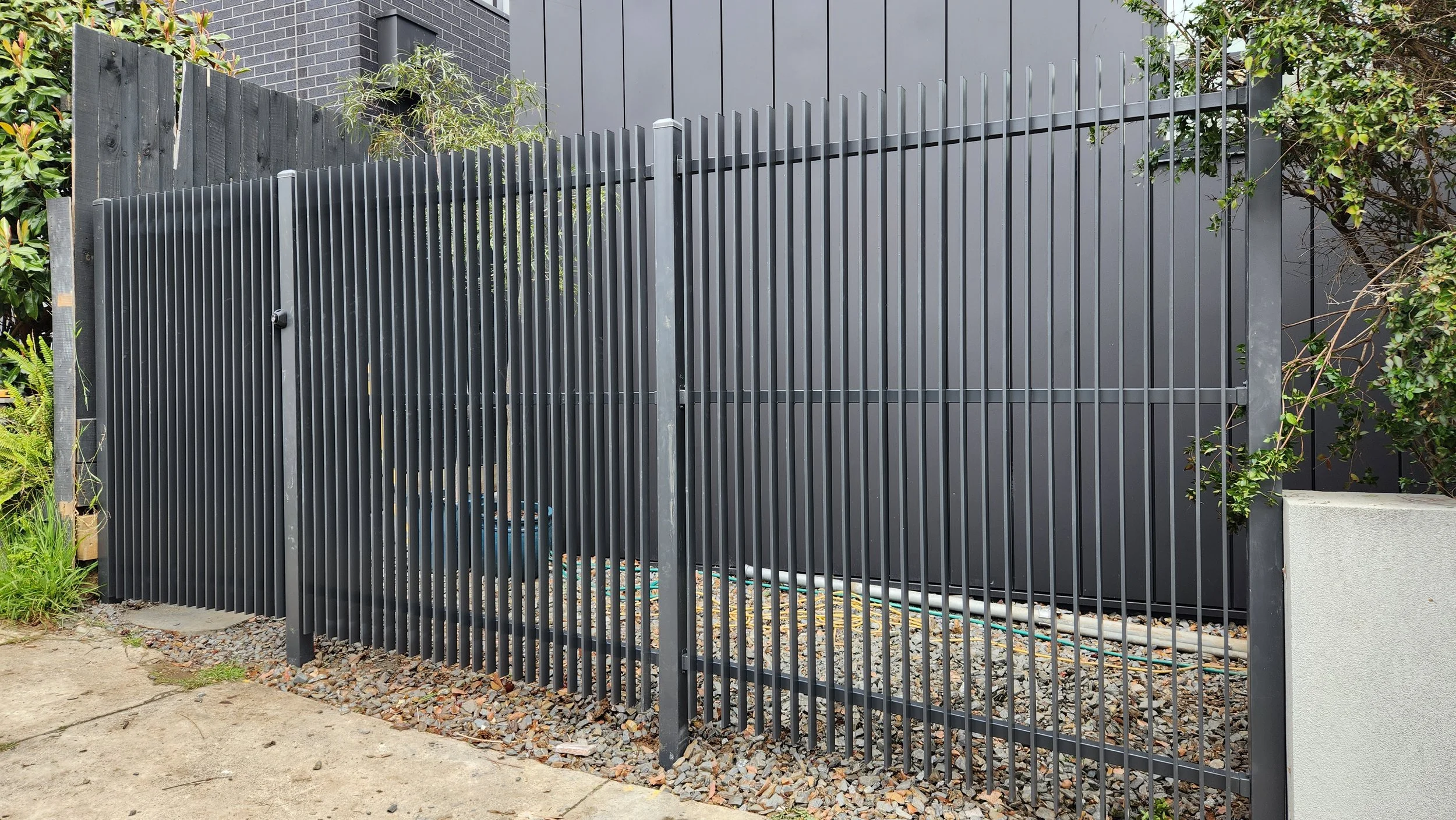 A black metal vertical slat fence with a sliding gate, surrounded by greenery, on a gravel and concrete ground.