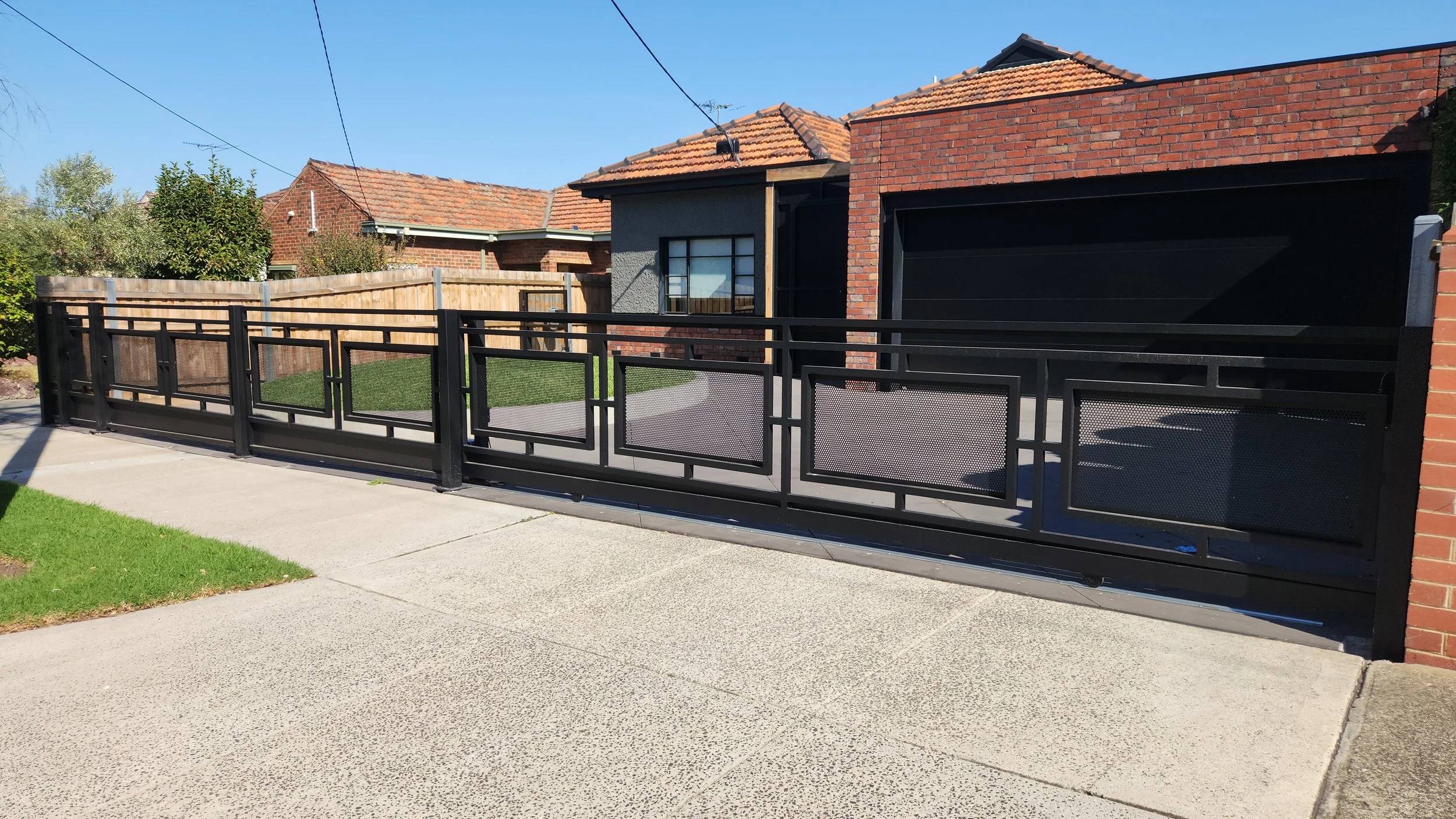 Modern black metal fence with panels and mesh design, installed in front of a brick house with gray and brick walls, red tile roof, and a small yard with grass, on a concrete sidewalk under a clear blue sky.