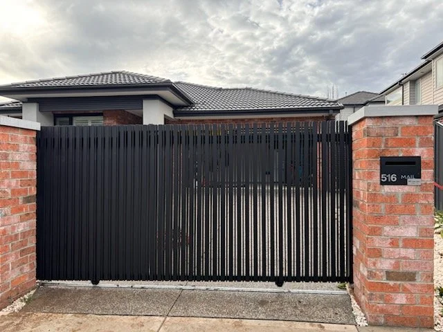 Black metal gate flanked by brick pillars with house and cloudy sky in the background.