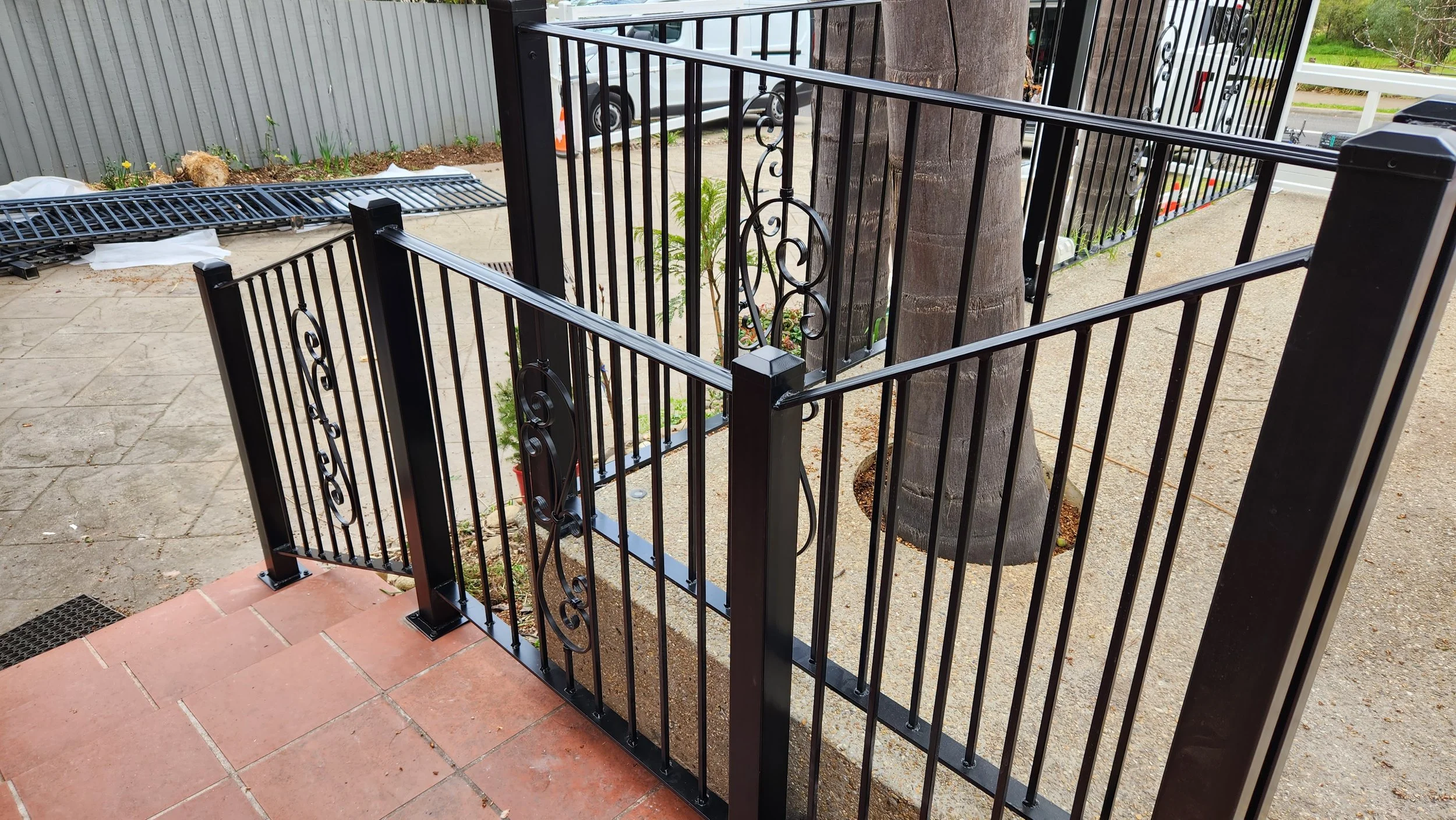 Black metal safety gate with decorative scrollwork installed on a porch with a concrete sidewalk and a tree in the background.