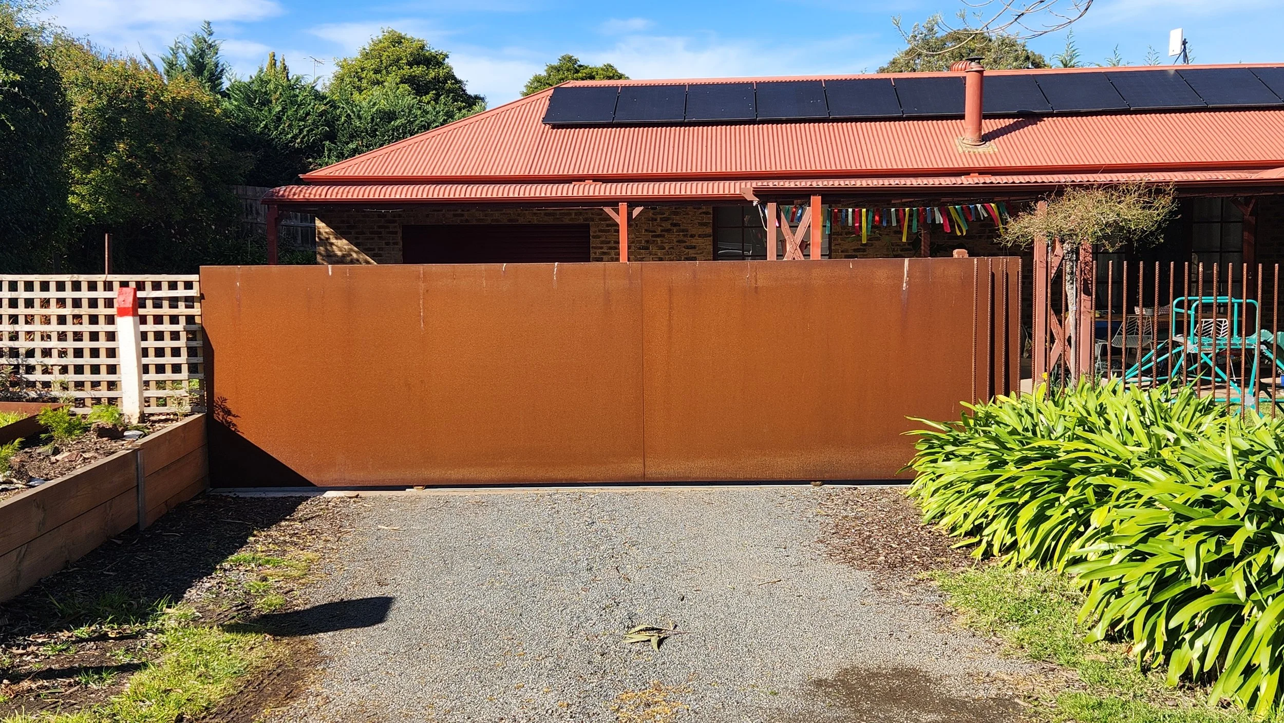 A large rust-colored metal gate in front of a house with a red metal roof, solar panels, and solar hot water system. Green shrubs and garden beds on either side, with clear blue skies above.