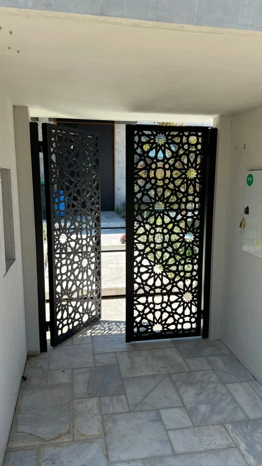 Decorative black metal gates with intricate geometric patterns, partially open, leading outside to a sunny area with steps and some greenery.