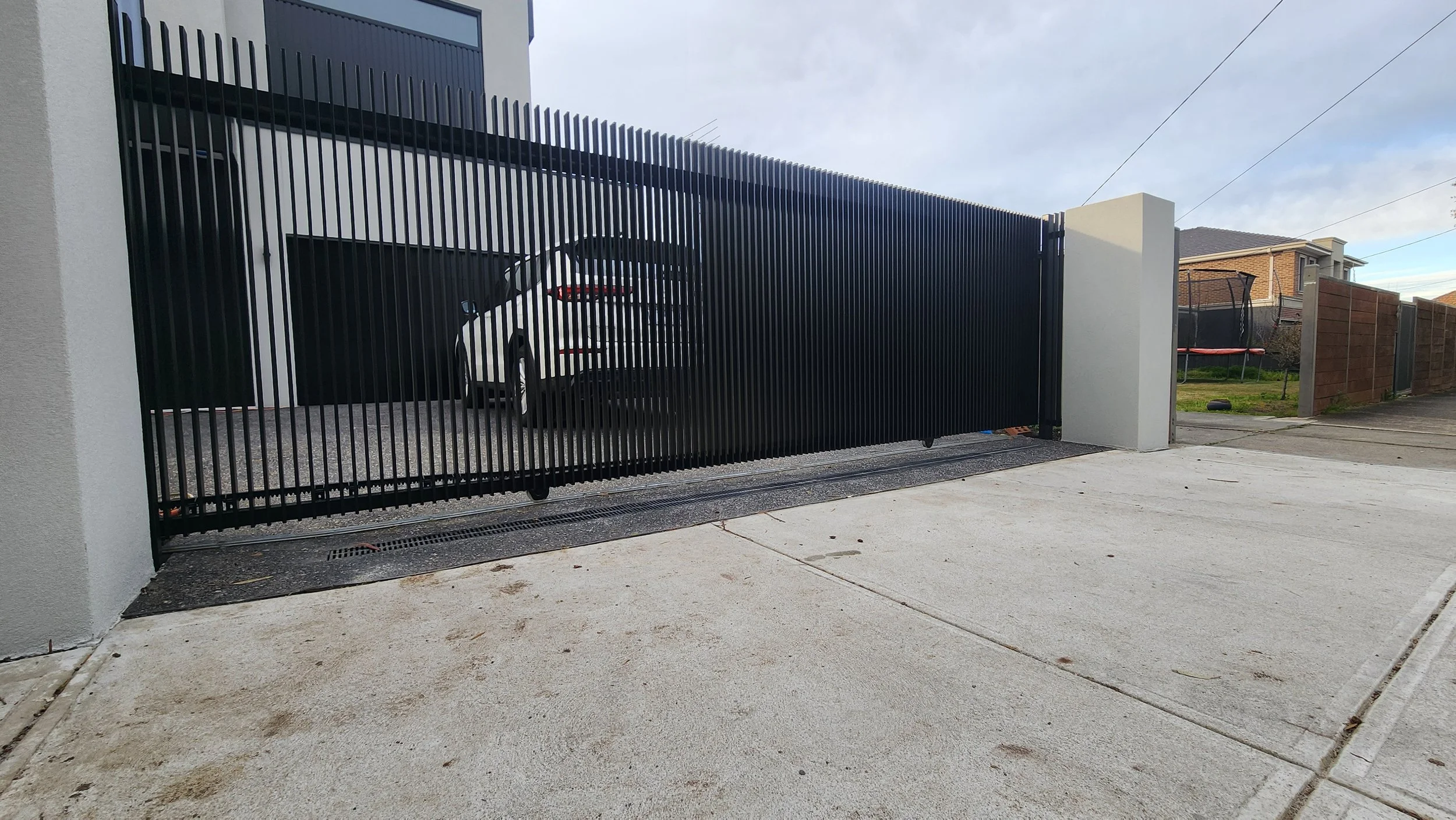 Black metal sliding gate open to reveal a white car parked in a driveway in front of a modern house.