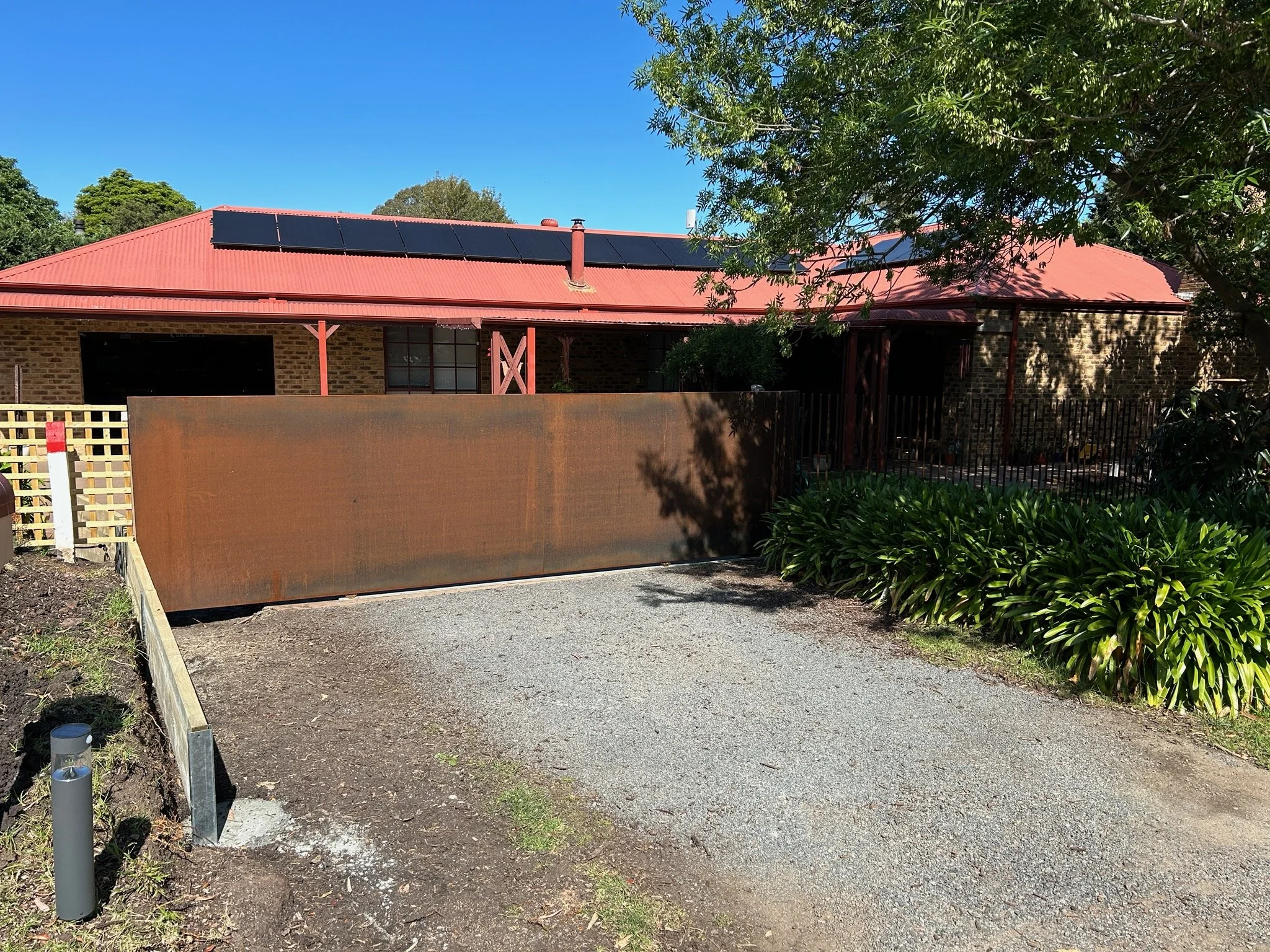 A house with a red roof and solar panels, brick walls, surrounded by green trees and plants, with a rust-colored gate in front.