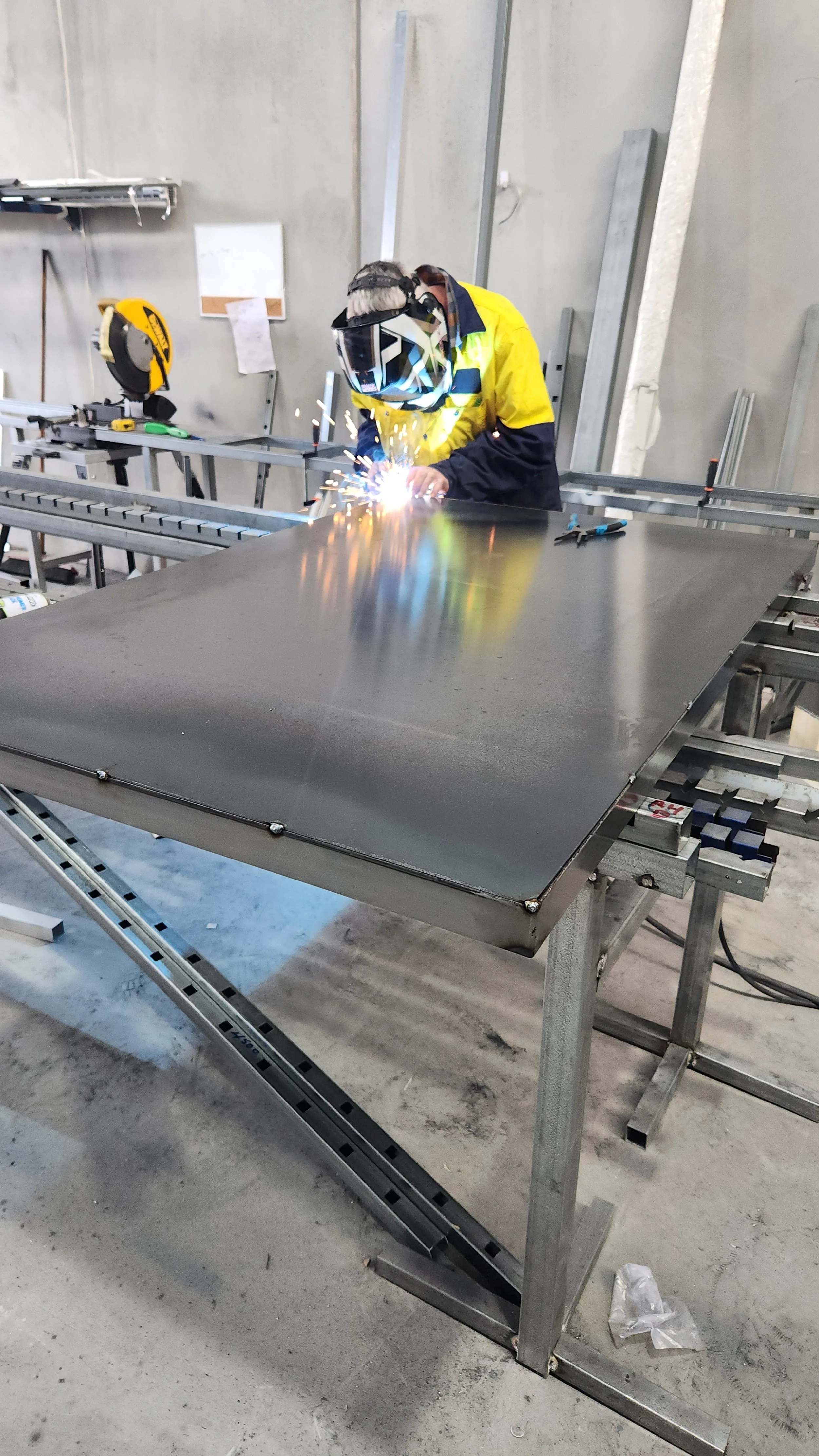 A person welding a metal sheet in an industrial workshop, wearing a protective helmet and yellow safety jacket.