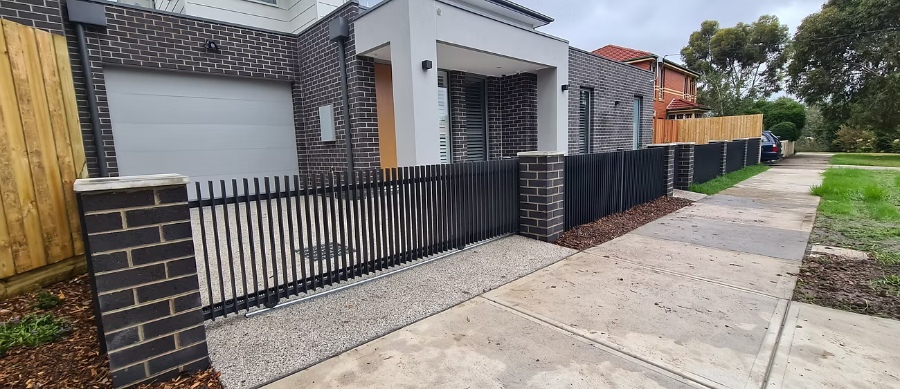 New modern house with black brick exterior, black metal fence, concrete driveway, and grassy yard on a cloudy day.