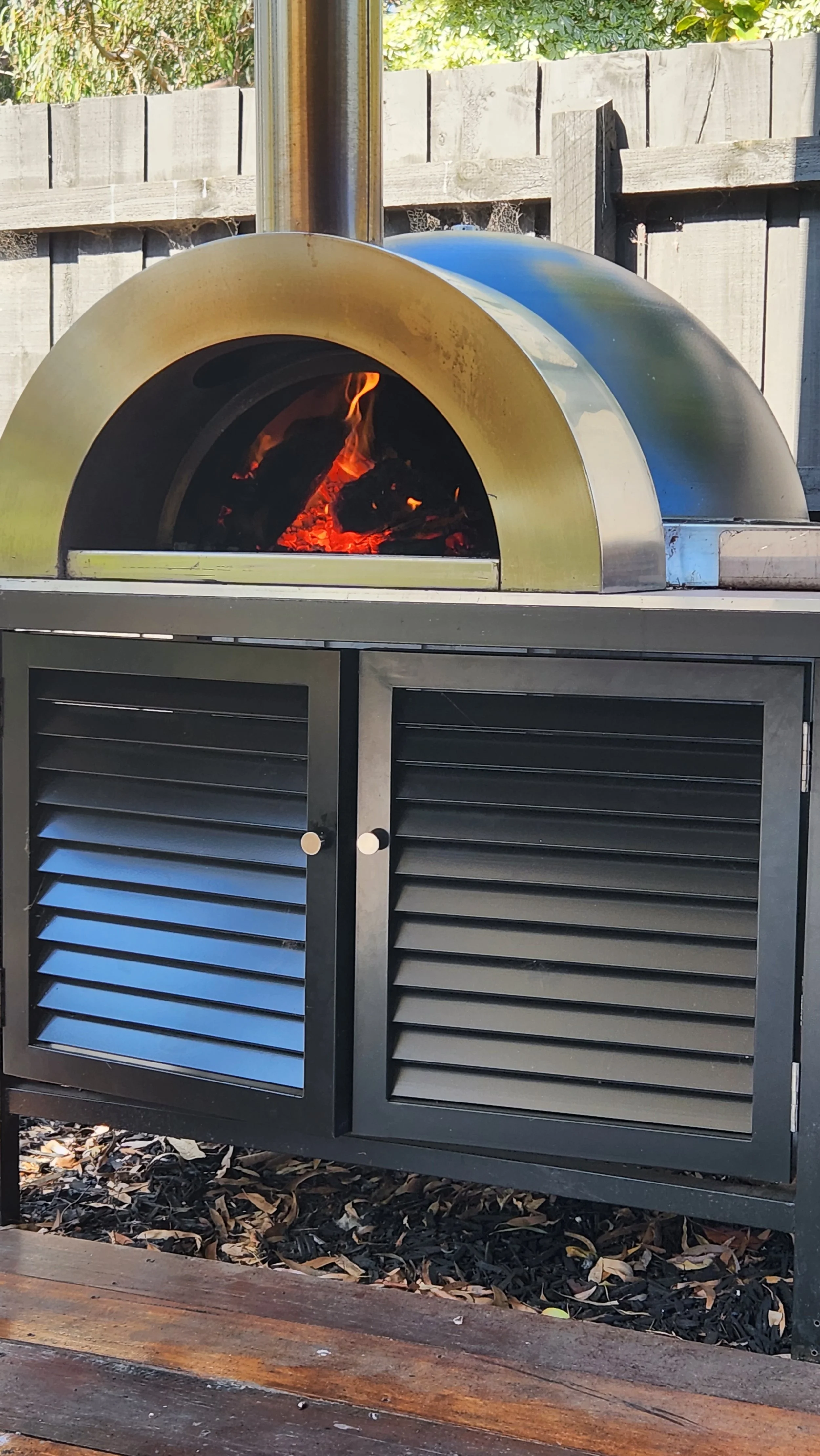 A modern outdoor pizza oven with a stainless steel dome and a burning fire inside, situated outside in a backyard with a wooden fence.