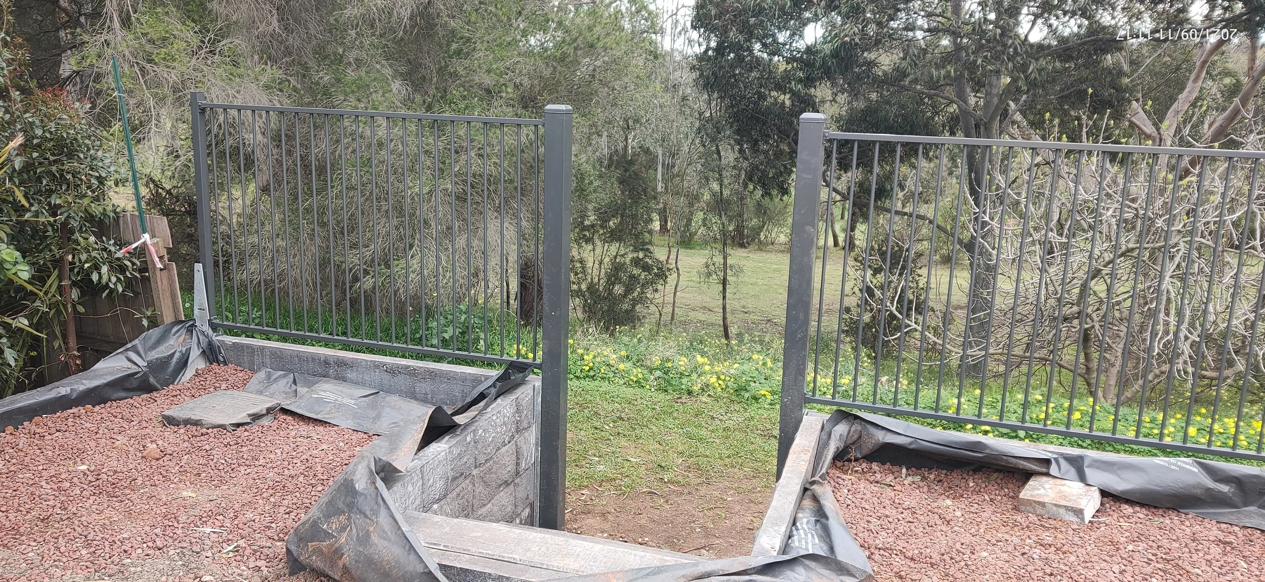 Partially constructed outdoor garden bed with black metal gate fence on a grassy area with trees and bushes in the background.