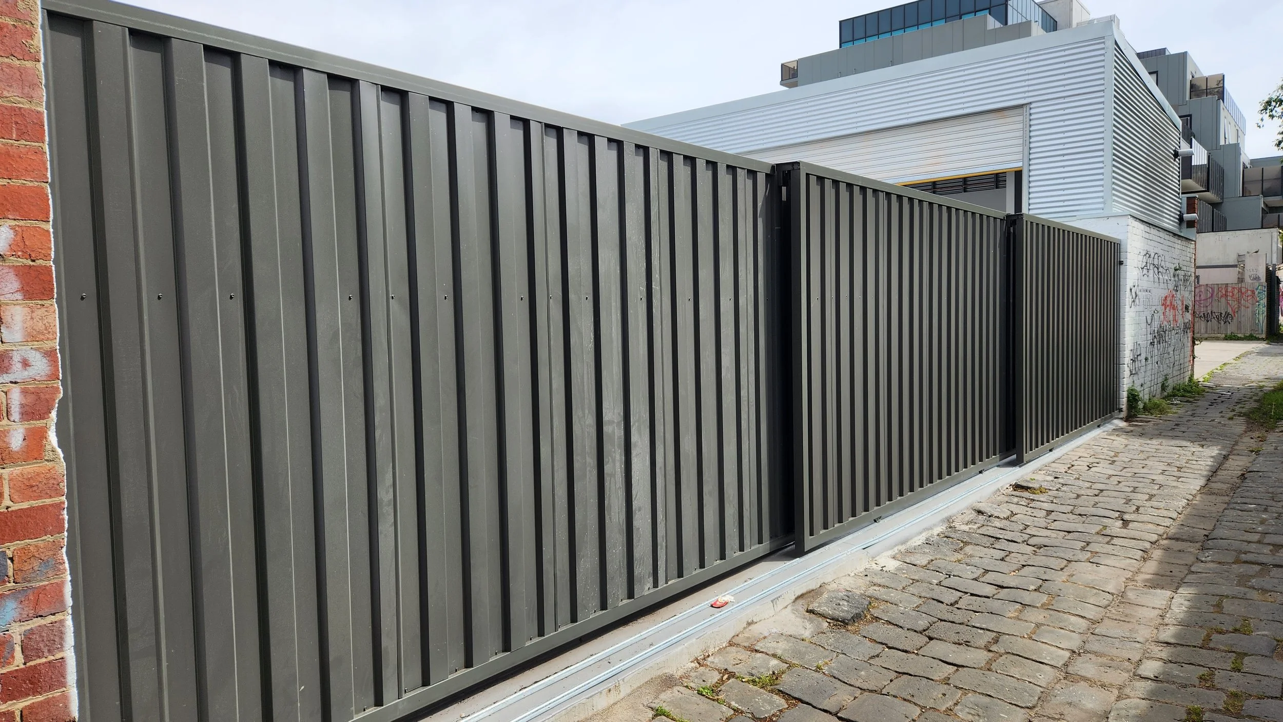 A tall black metal fence running along a brick sidewalk, with a brick wall partially visible on the left and a modern building in the background.