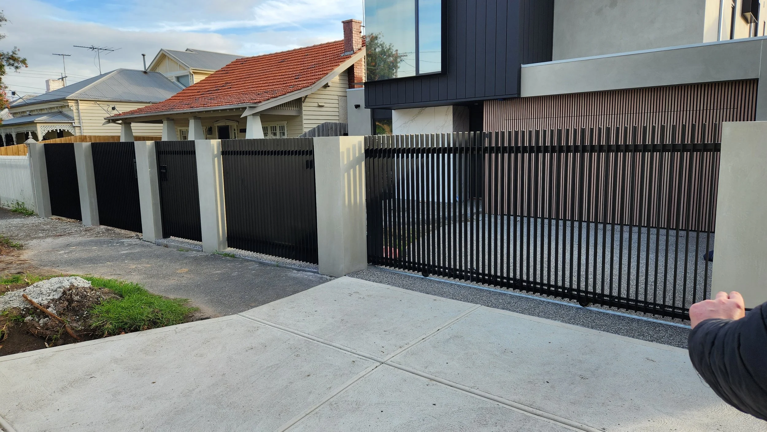 Modern black metal fence in front of residential houses, with a sidewalk in the foreground and a person's arm visible on the right side.