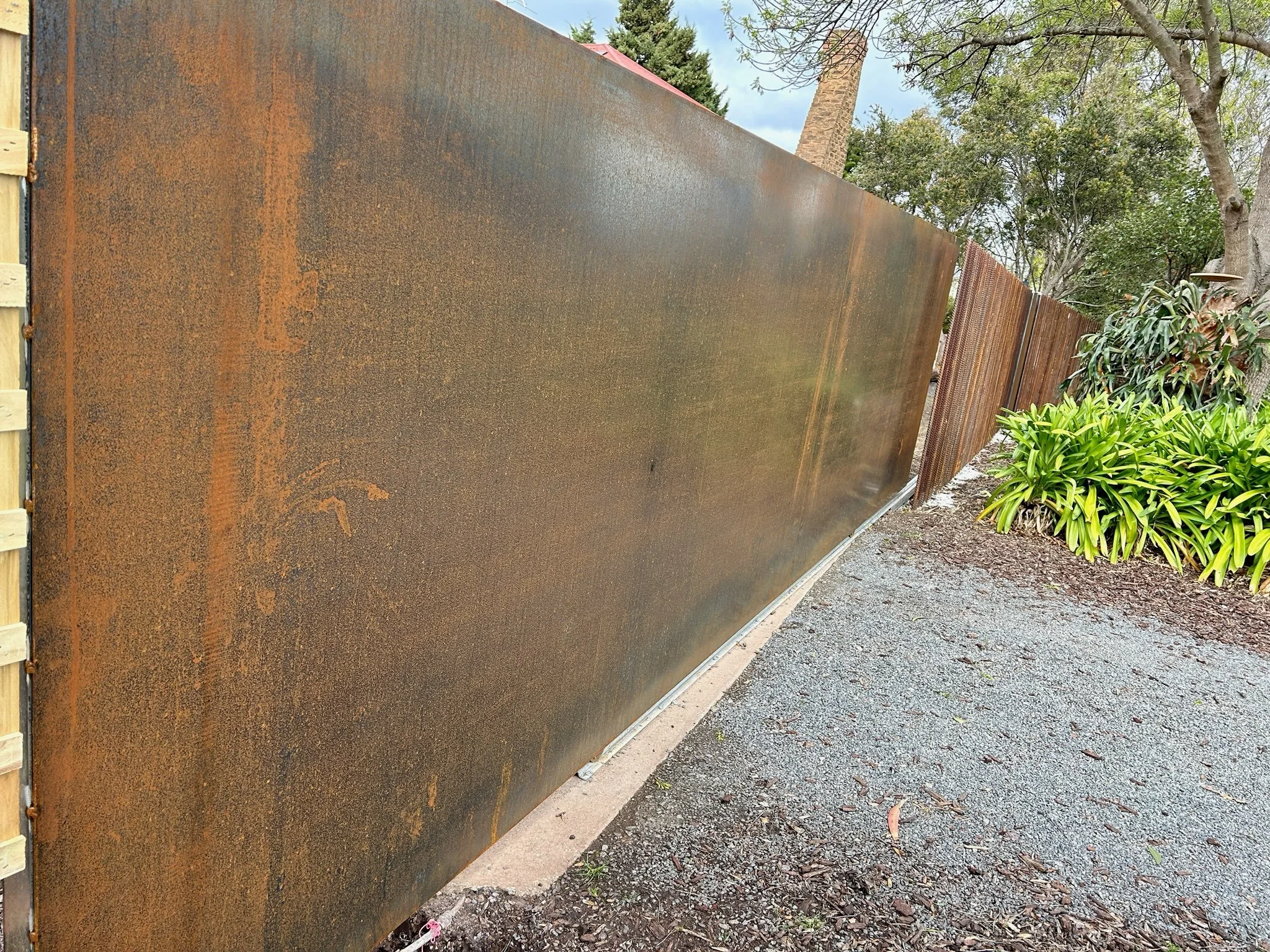 Watering trough or metal fence with rust on surface, beside a gravel path and green bushes, trees in background.