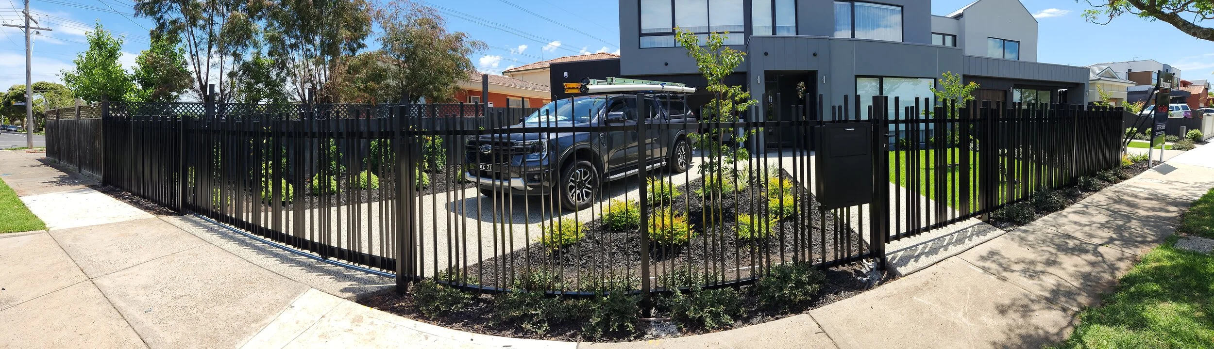 Modern residential building with a fenced parking area, containing a black pickup truck with roof racks, surrounded by landscaped garden, during sunny daytime.