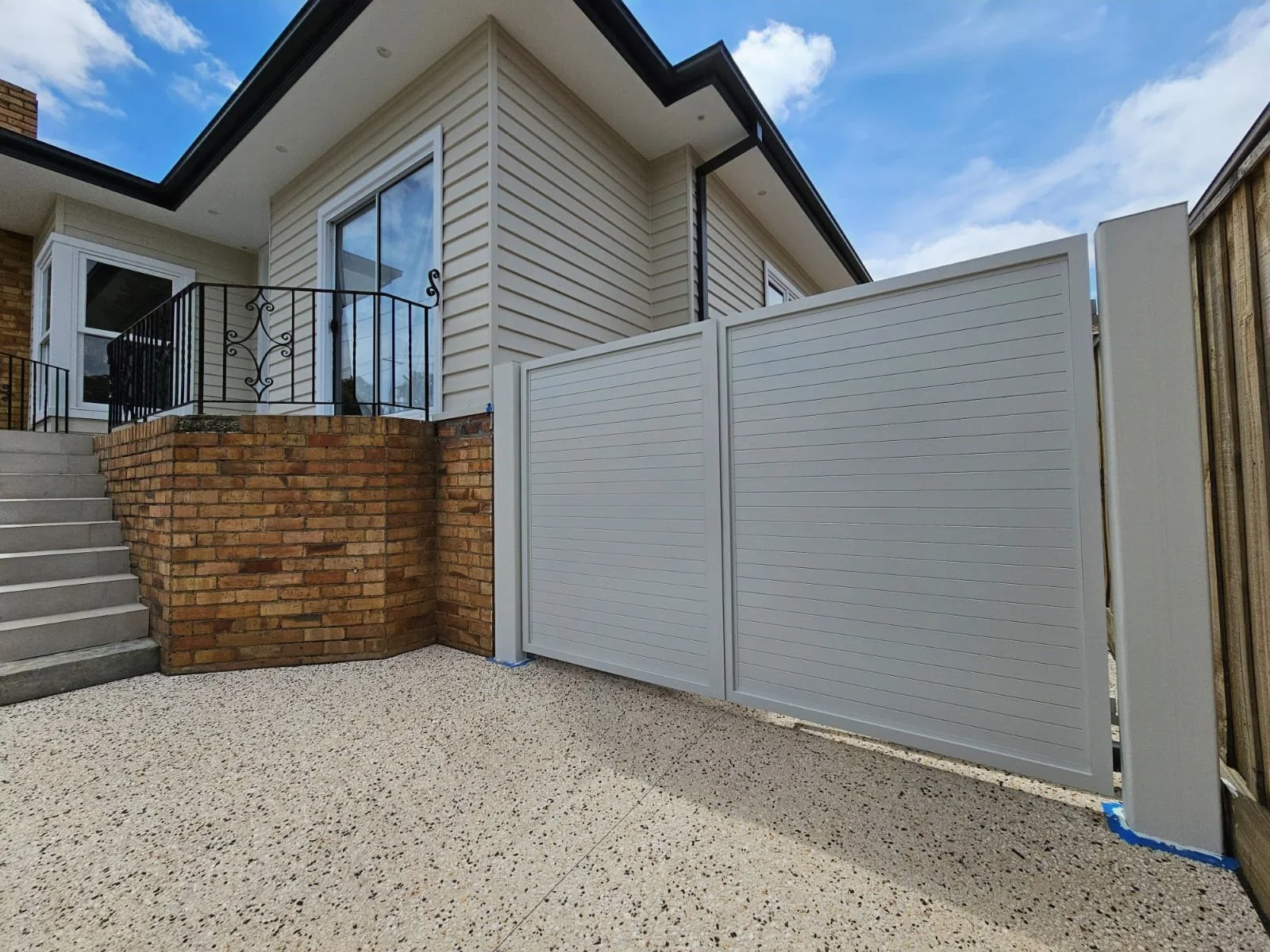 Modern house exterior with a brick foundation, white siding, metal balcony railing, concrete stairs, and a white privacy fence with a cloudy sky background.