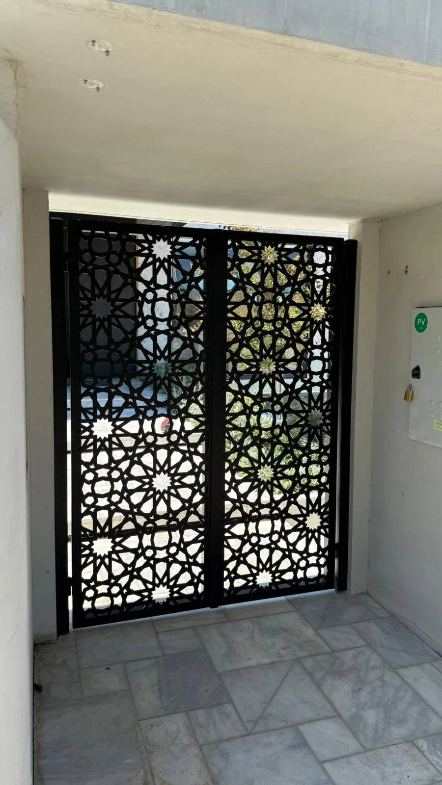 Intricate black metal gate with geometric and floral patterns at entrance of building, marble tiled floor, white walls, and utility box with green sticker on right side.