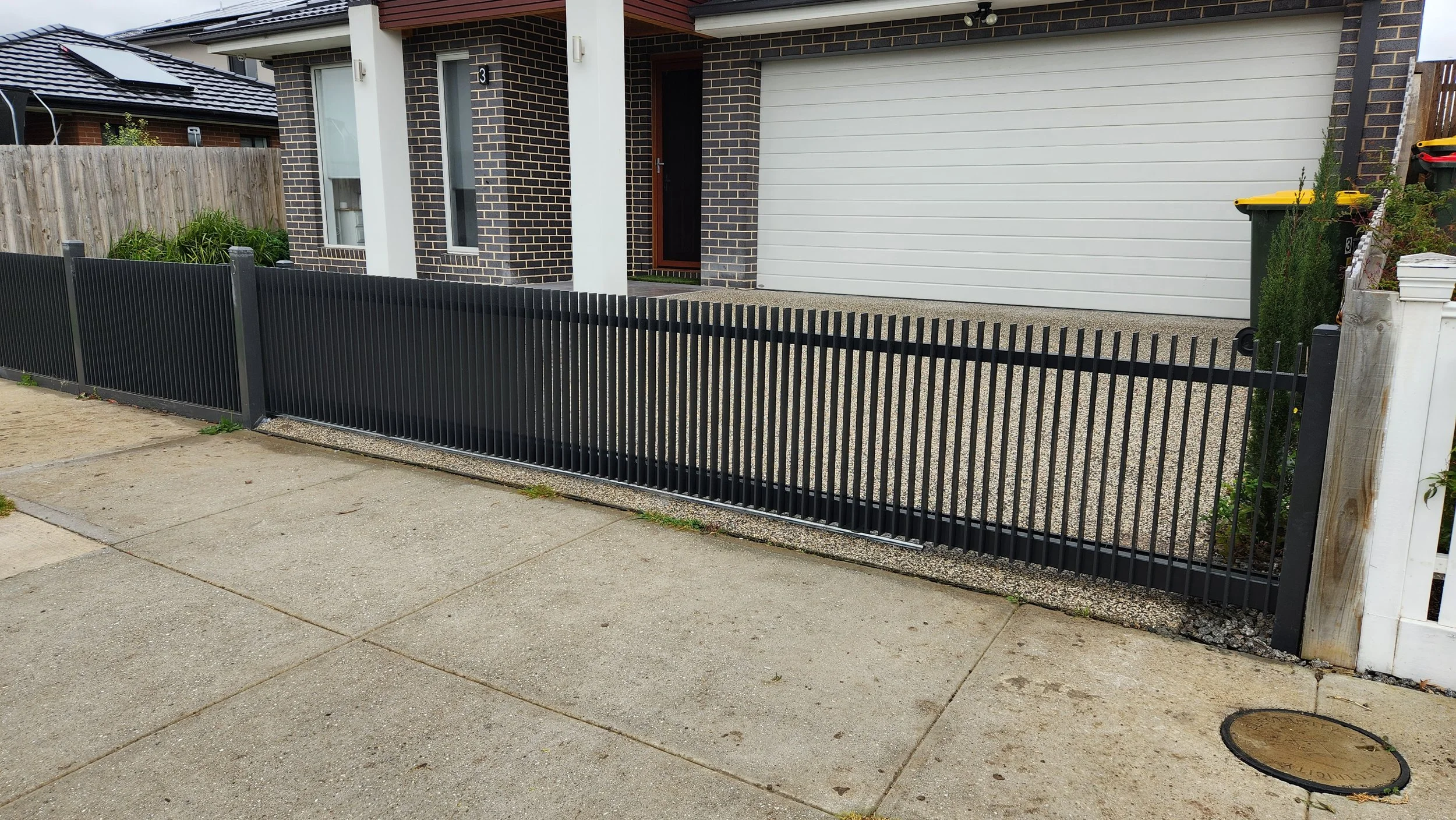 Modern house with brick facade, white garage door, concrete driveway, black metal fence, and partial view of the sidewalk and street.