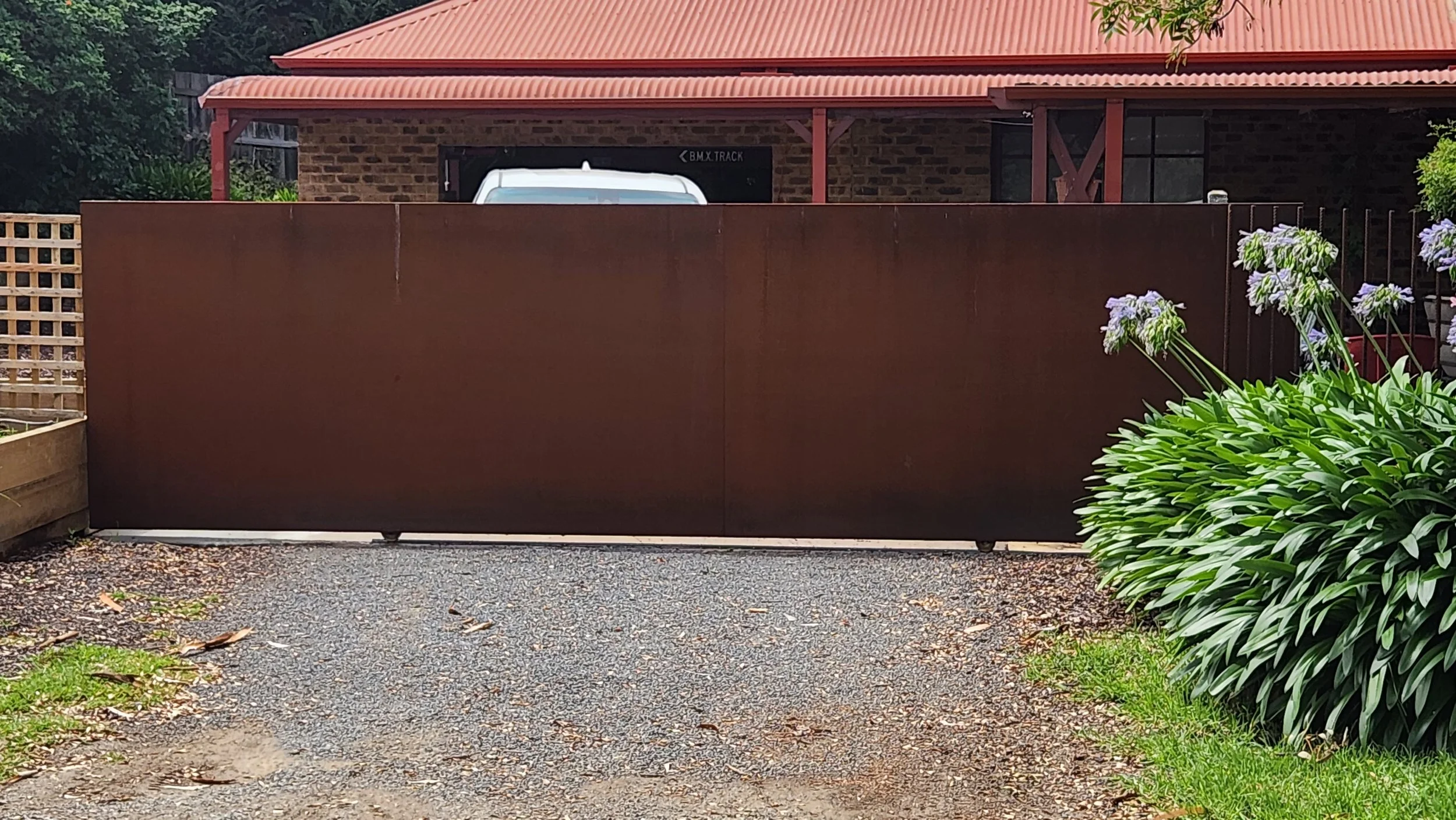 Brown sliding gate in front of a gravel driveway, with a large bush with purple flowers on the right and a house with a red roof in the background