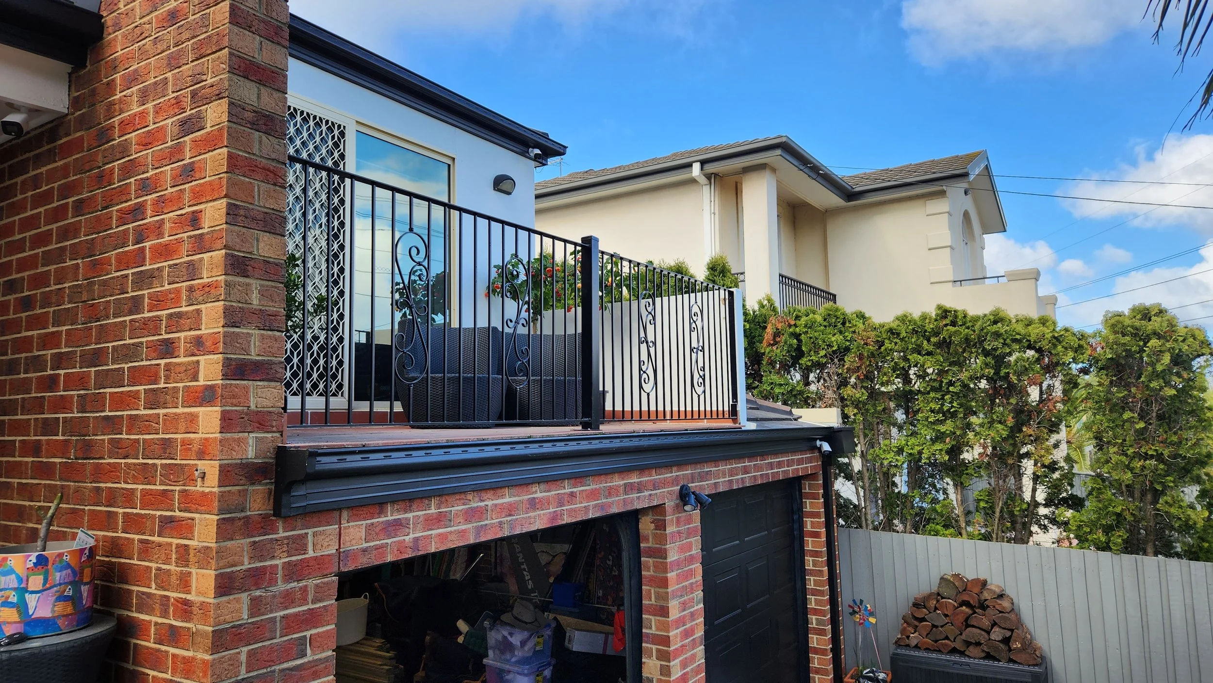 Backyard patio with black metal railing, potted plants, and outdoor wicker furniture, next to a house with white and brick exterior.