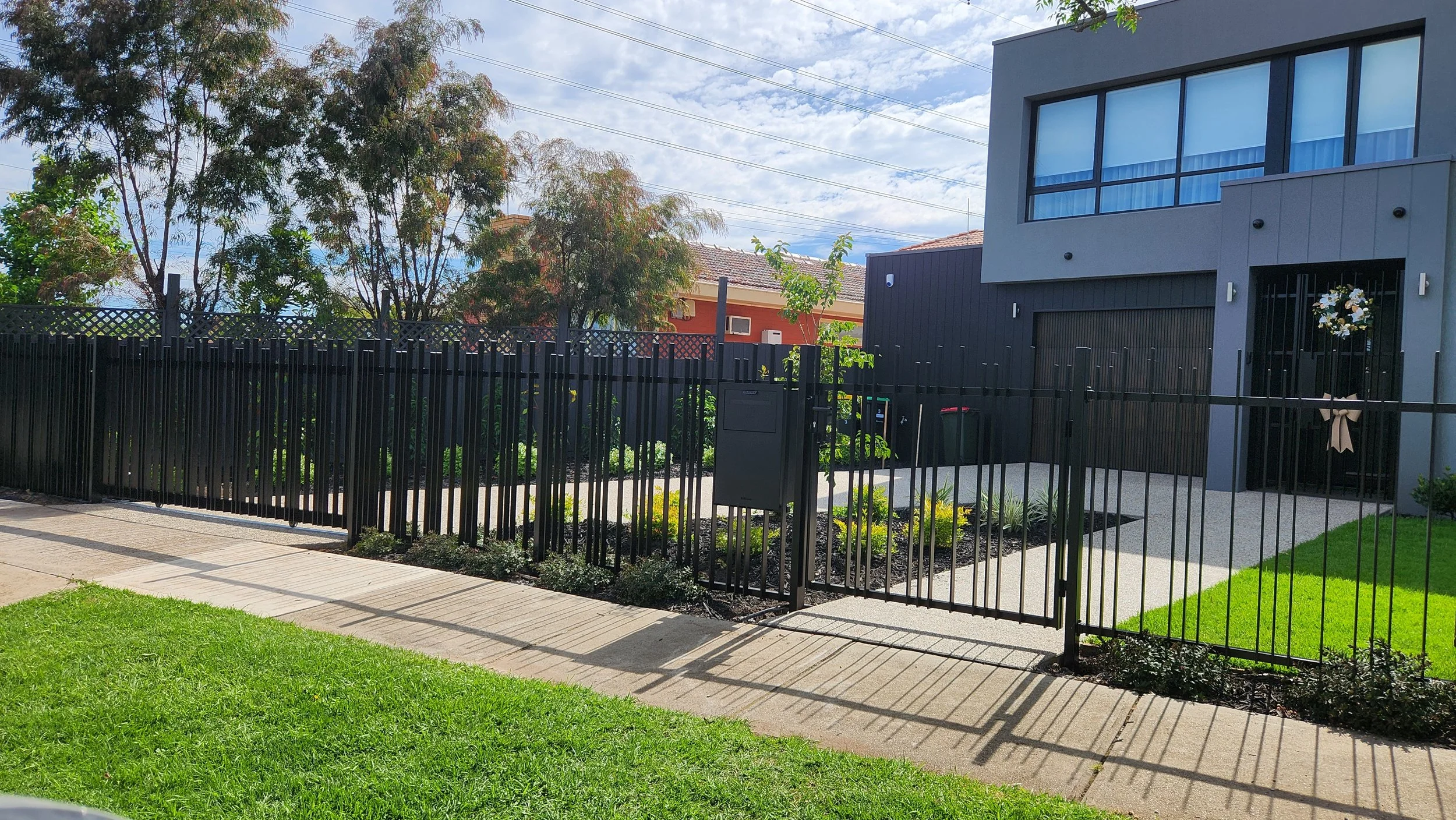 Modern gray house with large window and black garage door, surrounded by a black metal fence, green grass and garden, in a sunny neighborhood.
