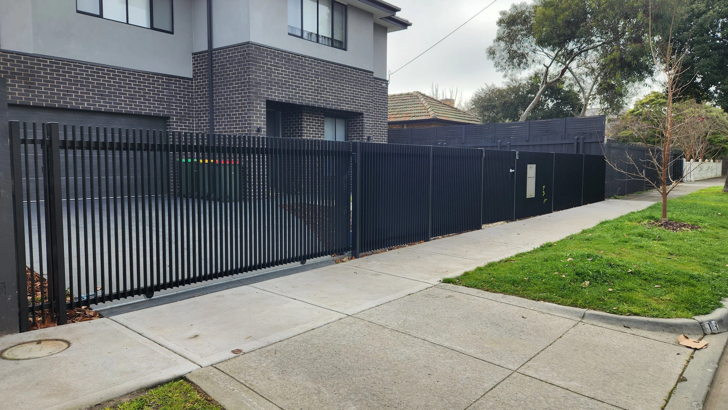 Black metal fence enclosing a residential driveway and yard, with a sidewalk and small grassy area with trees in front.