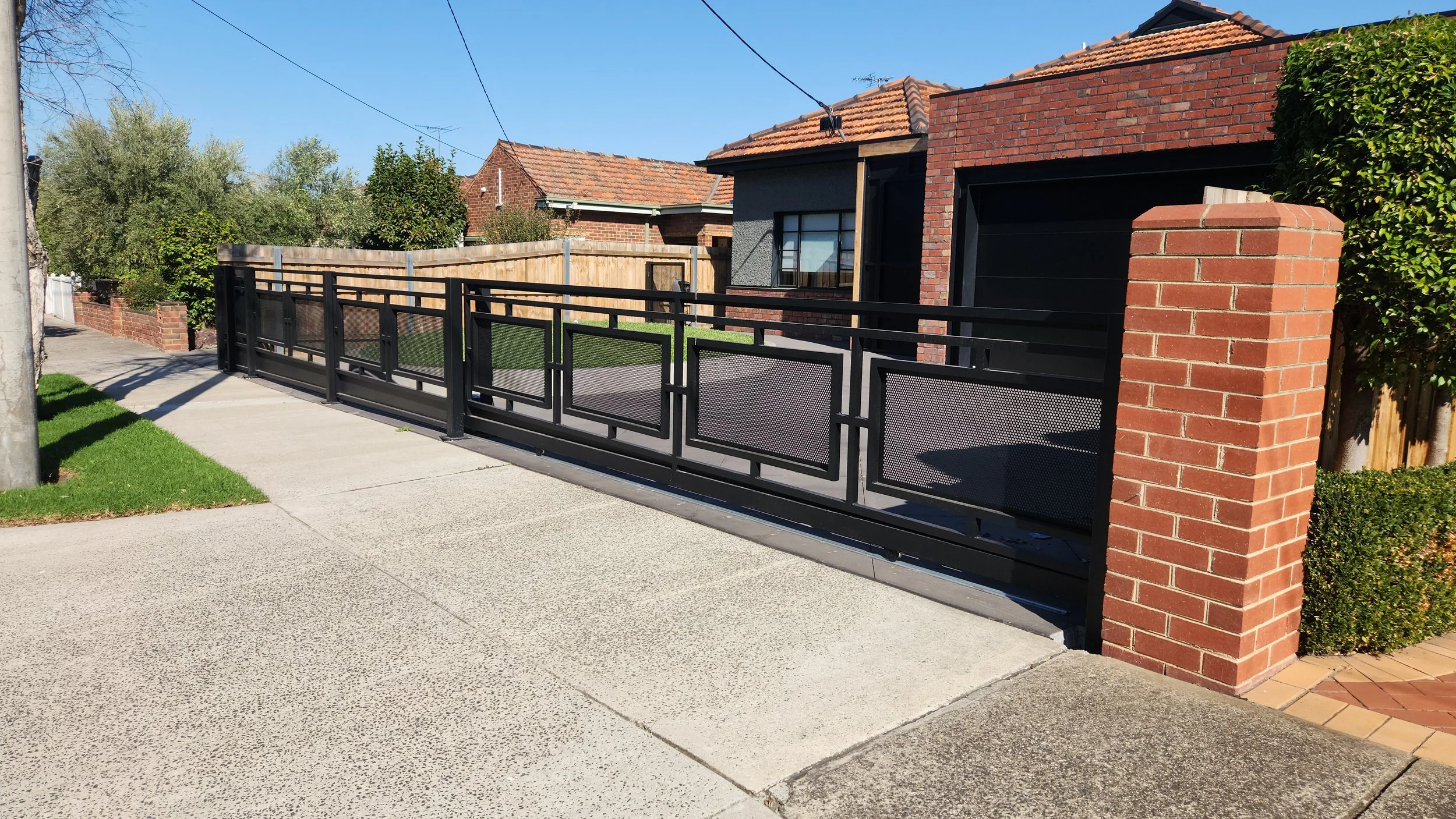 Black security gate with geometric design and mesh panels in front of a brick house and sidewalk.