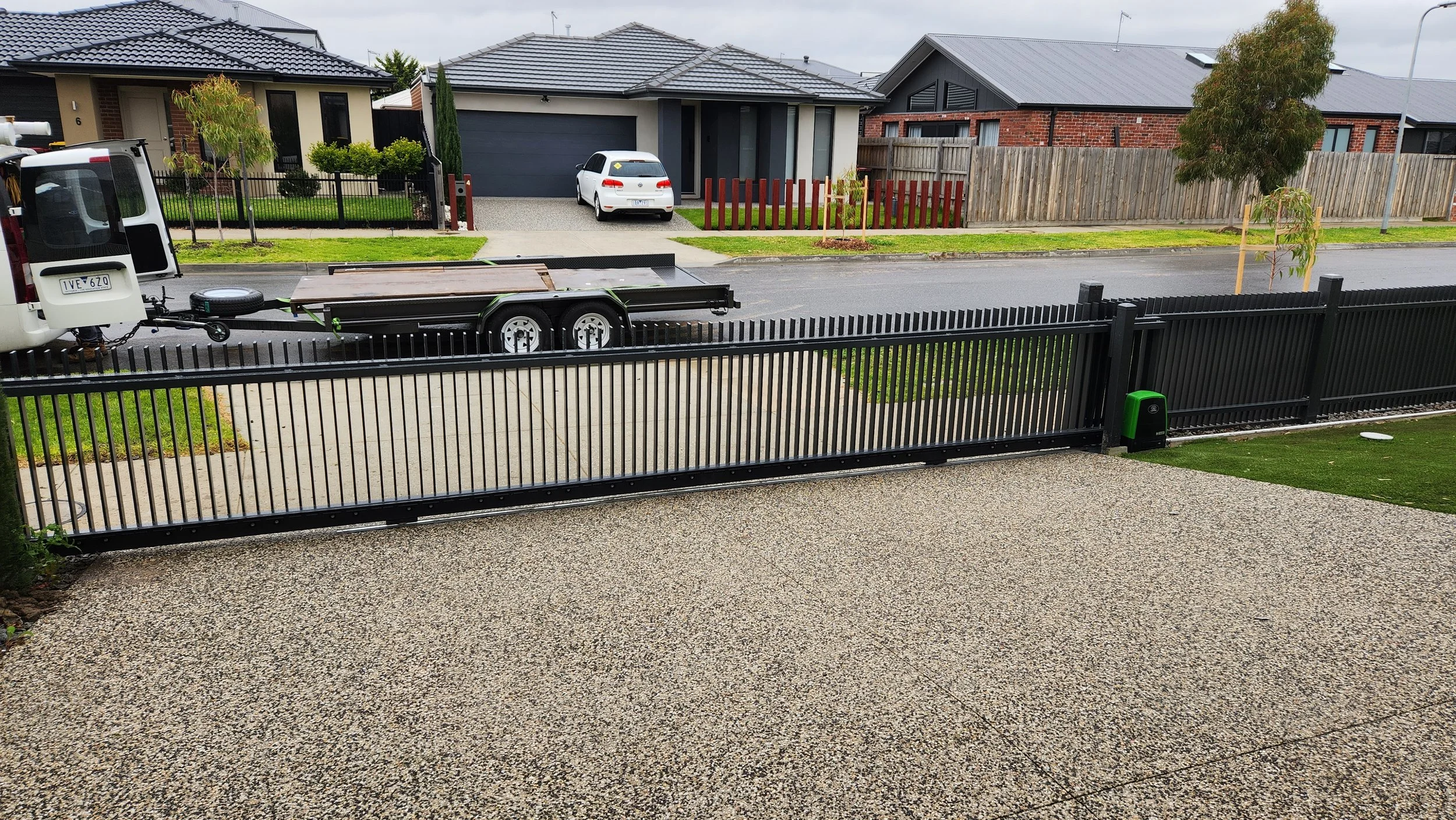 View of a suburban street with houses, a driveway, a white car, and a black metal sliding gate in the foreground.