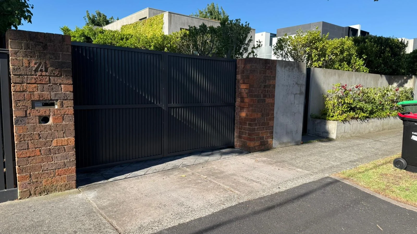Black metal gate with brick pillars, a white concrete wall to the right, plants and shrubs behind, and a black trash bin with red and green lids outside on the sidewalk.