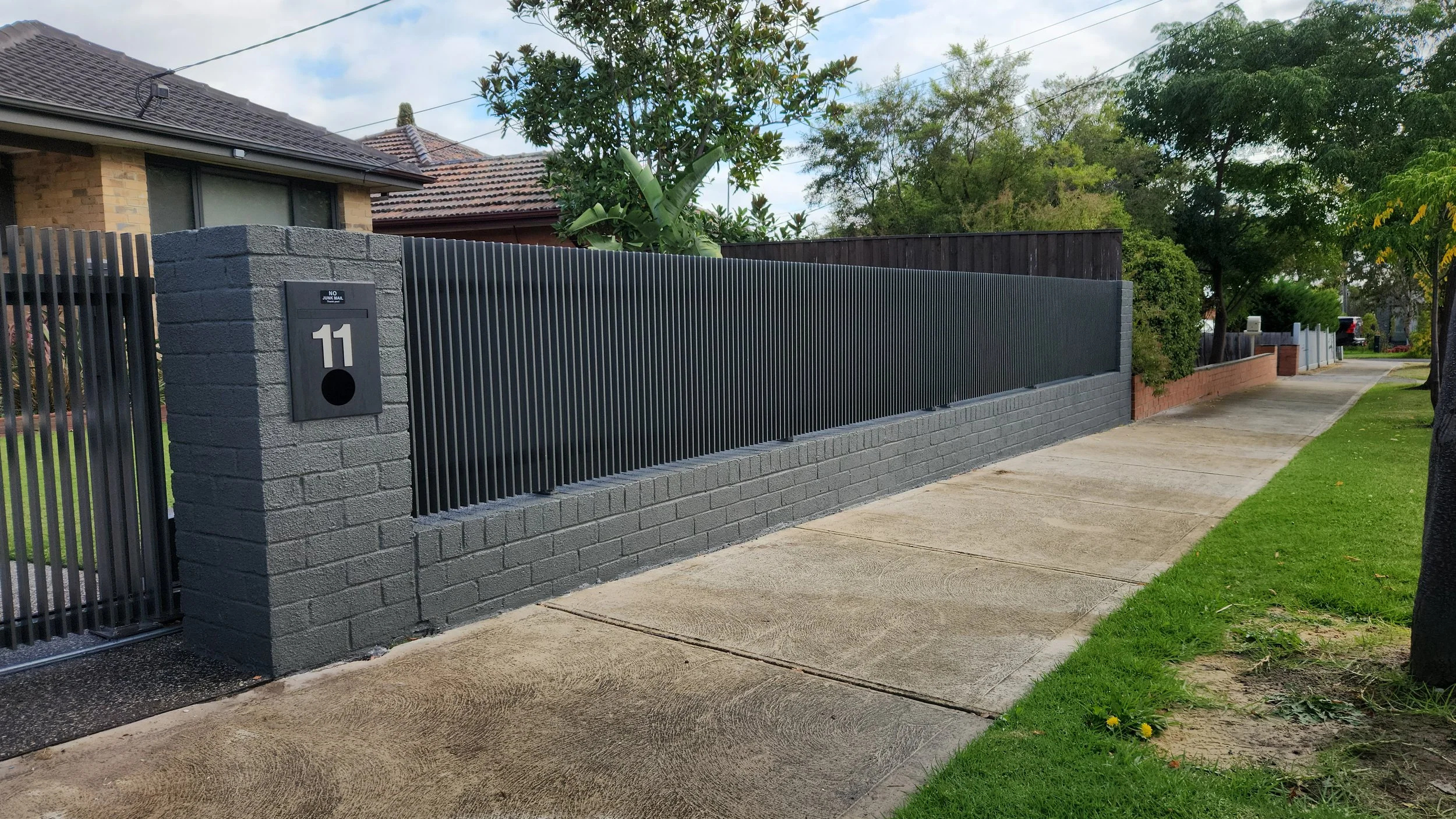 Street view of a modern black metal fence on a brick wall, with house number 11 and a mailbox, sidewalk, and grassy lawn with trees nearby.