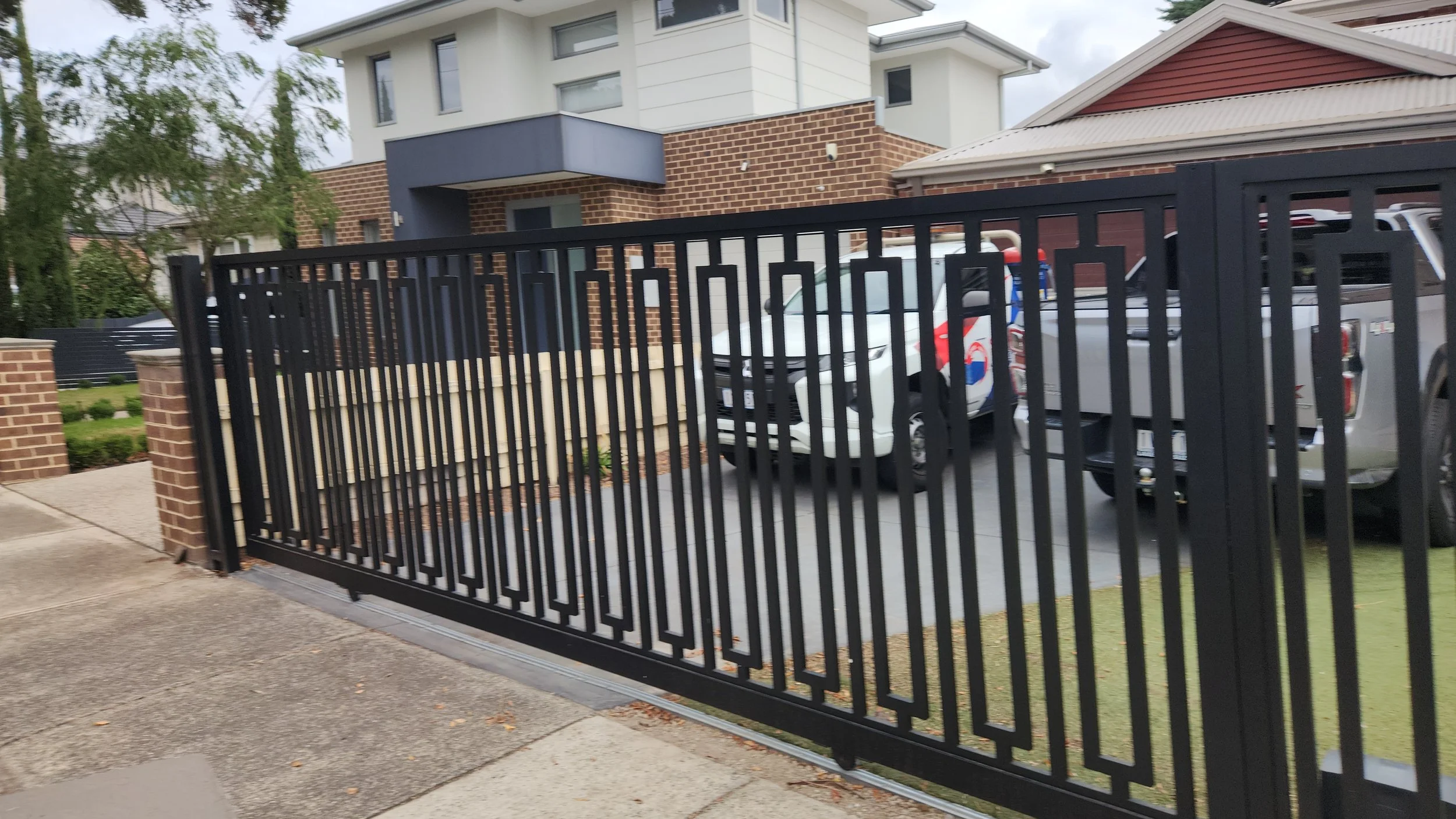 Black metal sliding gate in front of a residential driveway, with a townhouse and parked cars in the background.