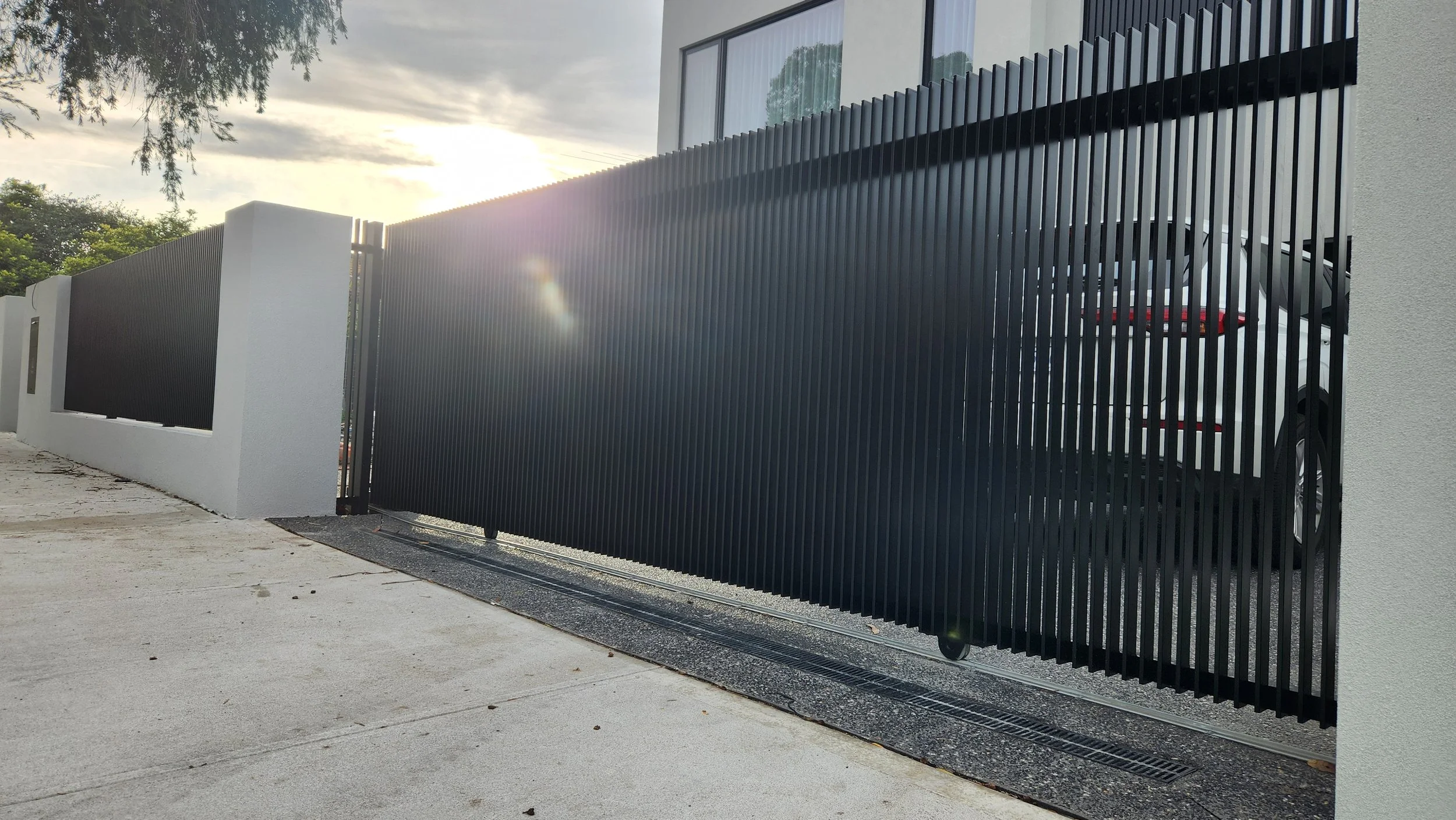 Modern black vertical slat gate with a sliding mechanism, adjacent to a white wall, at the entrance of a contemporary home with a car parked inside