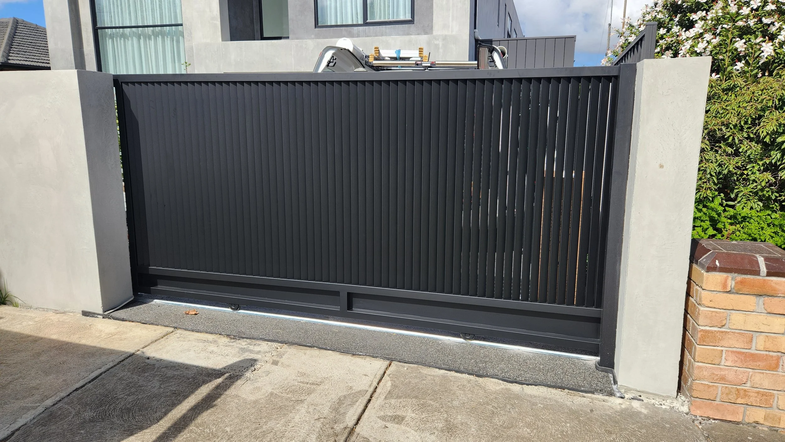 Black metal sliding gate with vertical slats, installed between concrete pillars, on a concrete driveway.