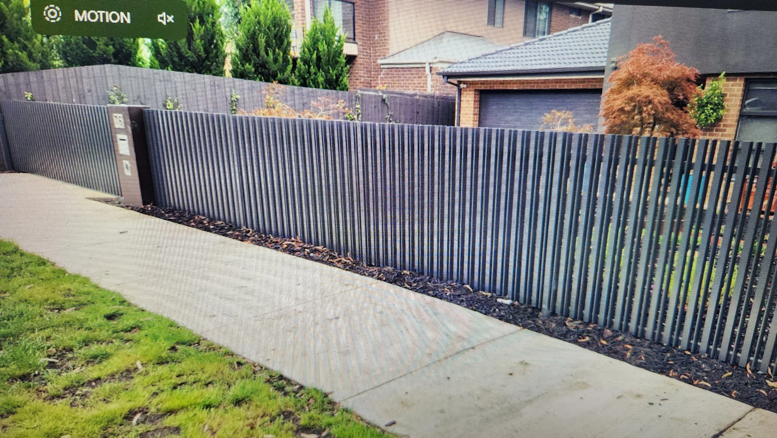 Residential backyard with a concrete sidewalk, a black metal fence, a brick house with a garage, and trees with autumn-colored leaves.