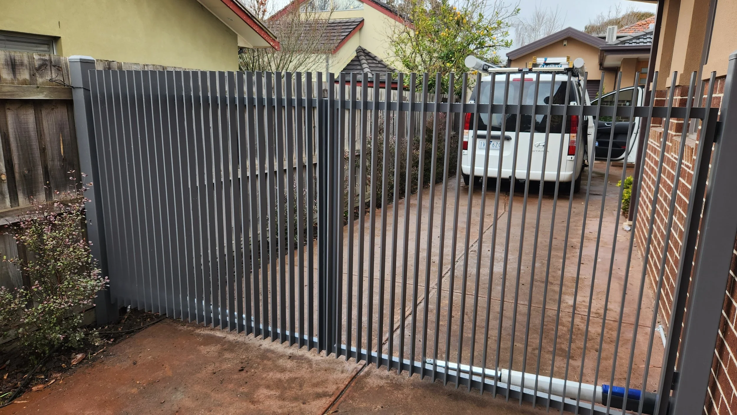 A gray metal sliding gate in front of a driveway with a white van parked inside, brick and wooden fencing on the sides, and residential houses in the background.