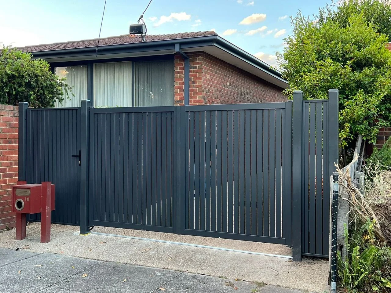 Black metal sliding gate in front of a brick house with a window and a hedge. Red mailbox on a wooden stand nearby.